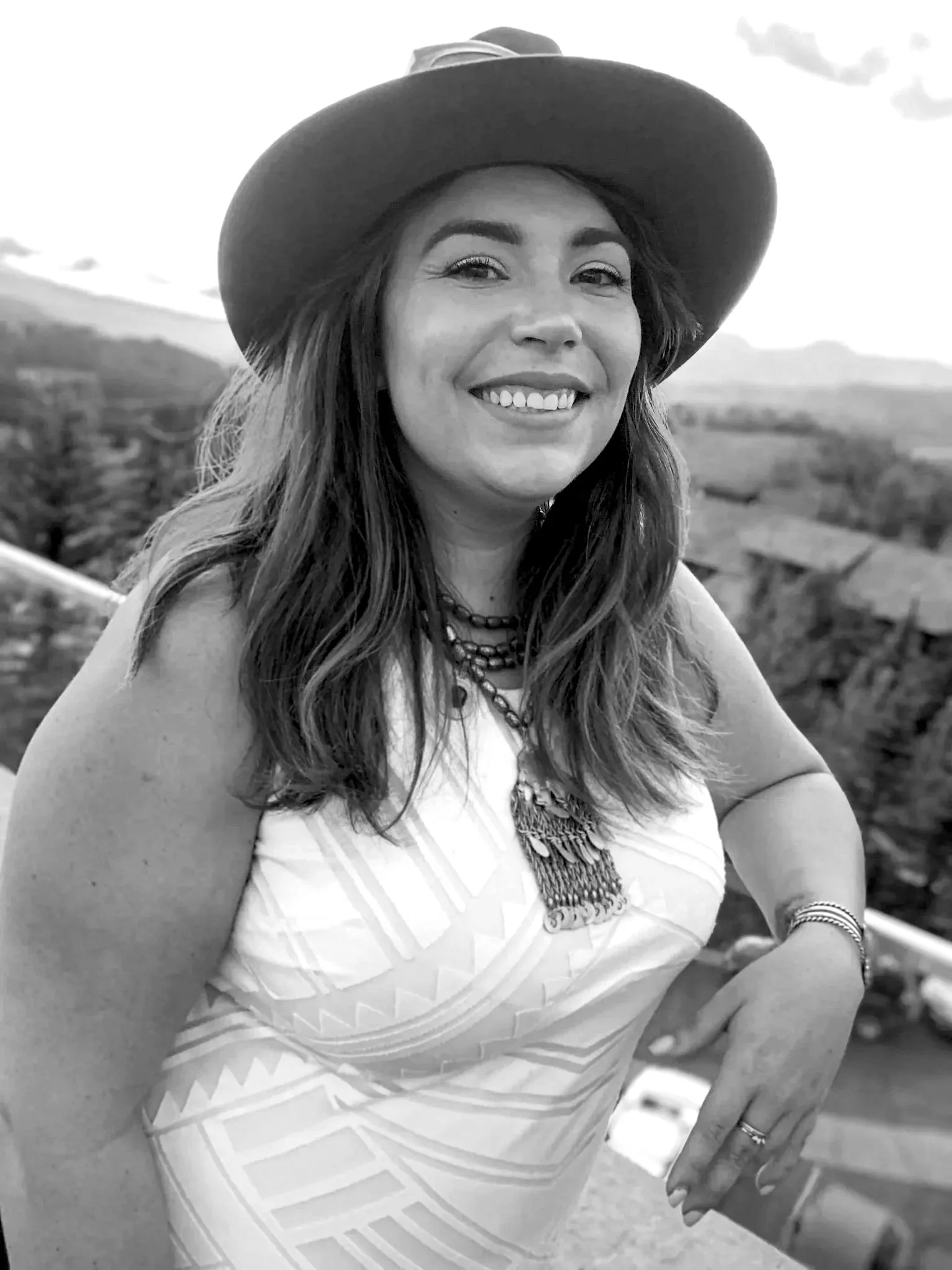 A woman smiling outdoors, wearing a large hat, patterned sleeveless top, multiple necklaces, and jewelry on her left wrist and finger, with a scenic landscape of trees and mountains in the background.