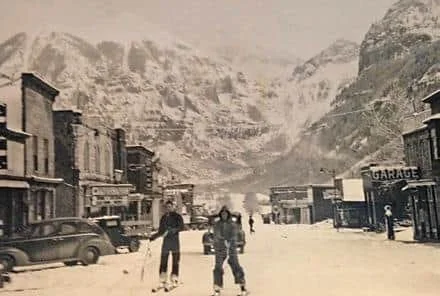 A snowy street scene in a mountain town with people walking, parked cars, and buildings, including a garage.