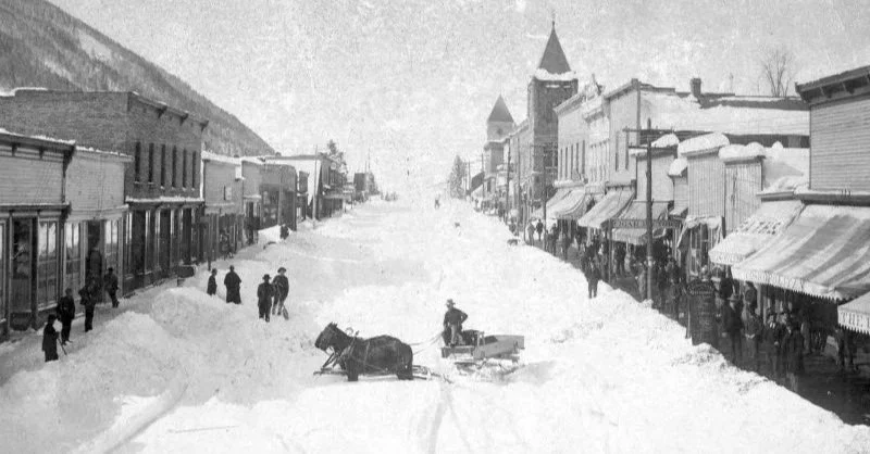 Historical black and white photo of a snow-covered main street in a small town, with buildings on both sides, horse-drawn sleighs, and several pedestrians.