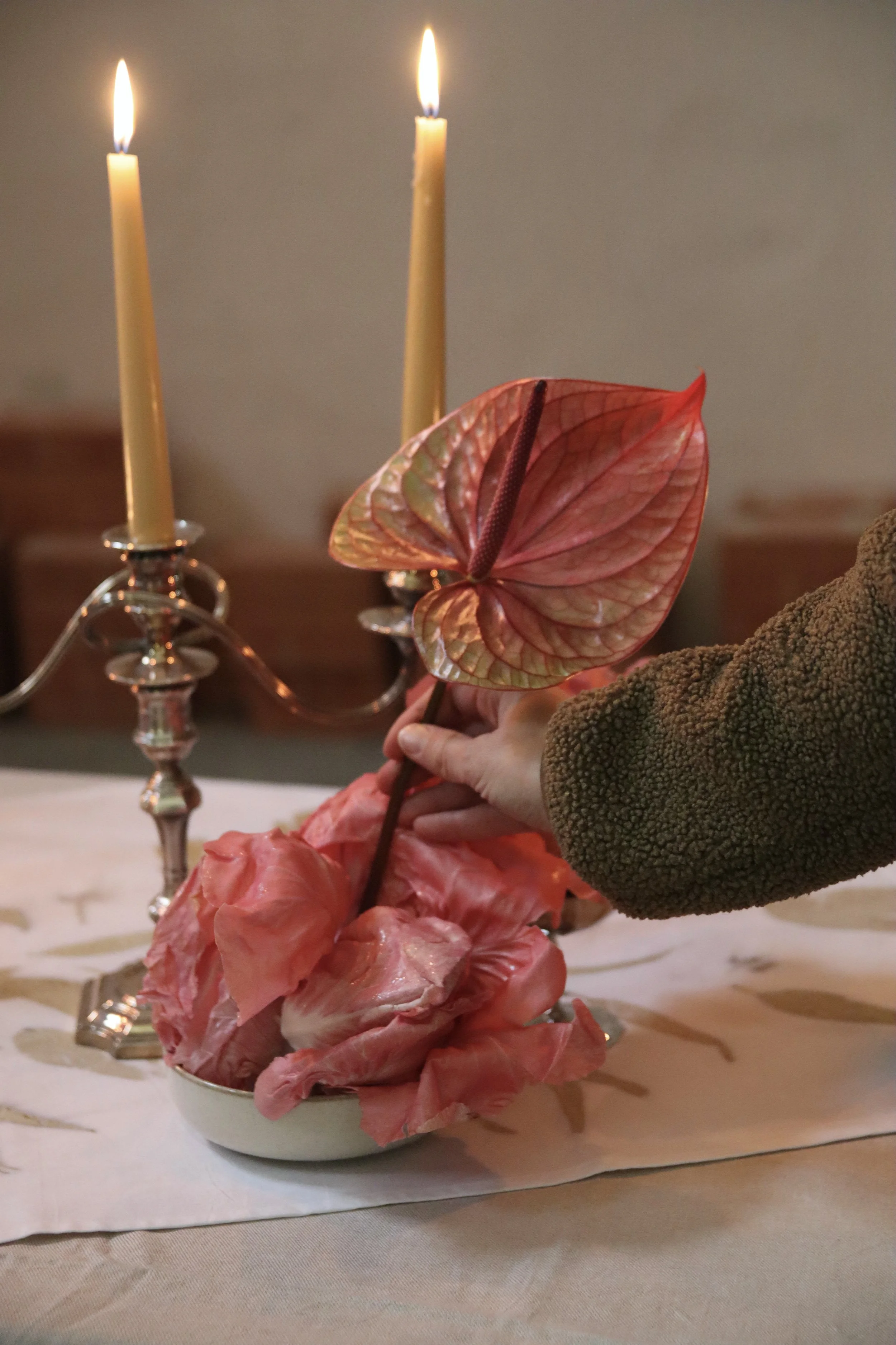 Una mano di una persona che tiene una foglia rossa, con due candele accese sullo sfondo e petali di fiori rosa in una ciotola.