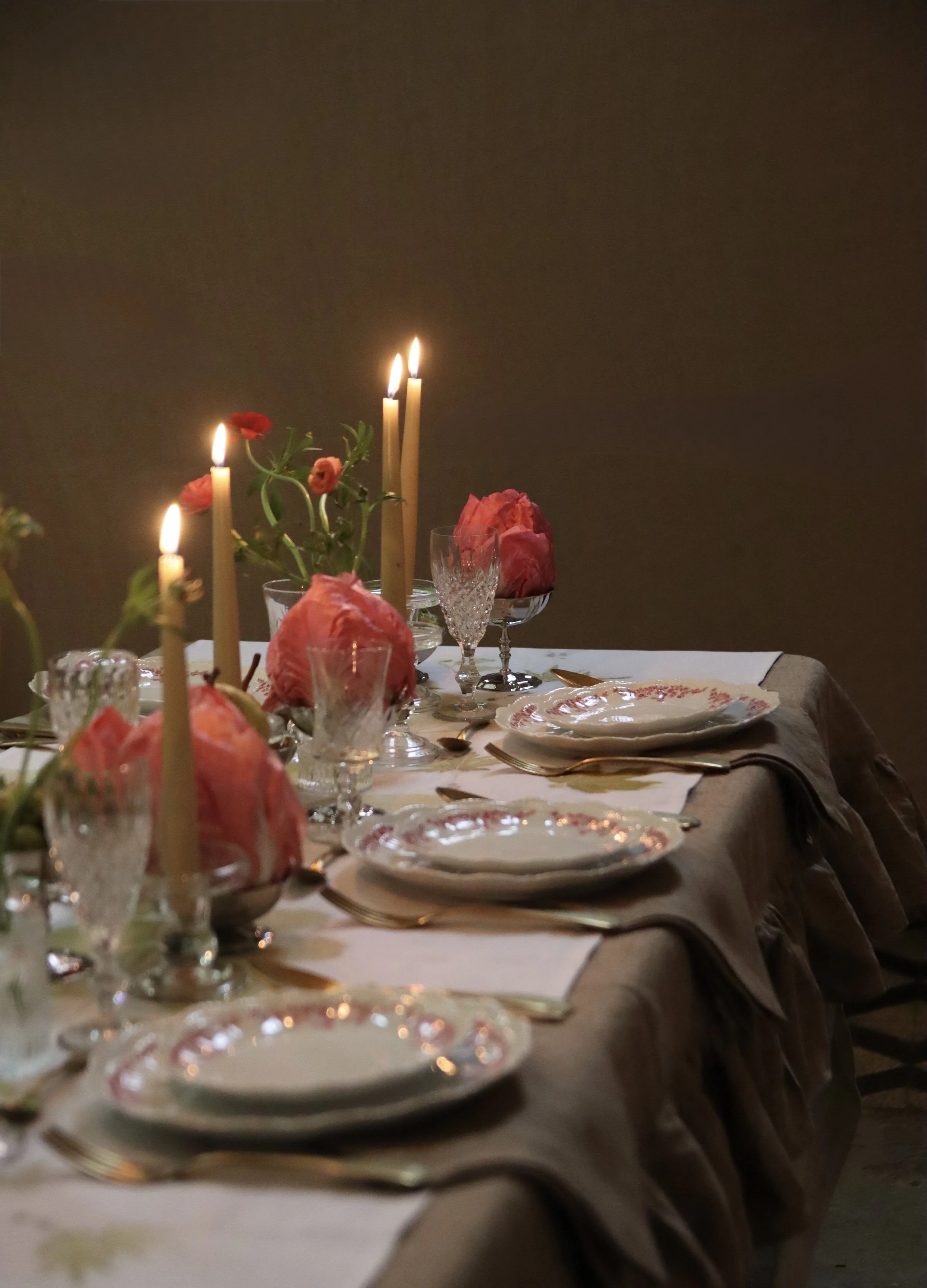 Una lunga tavola apparecchiata con piatti, bicchieri e posate, decorata con fiorellini e vasi, con candele accese e un grande fiore rosa al centro, in un'atmosfera elegante e intima.