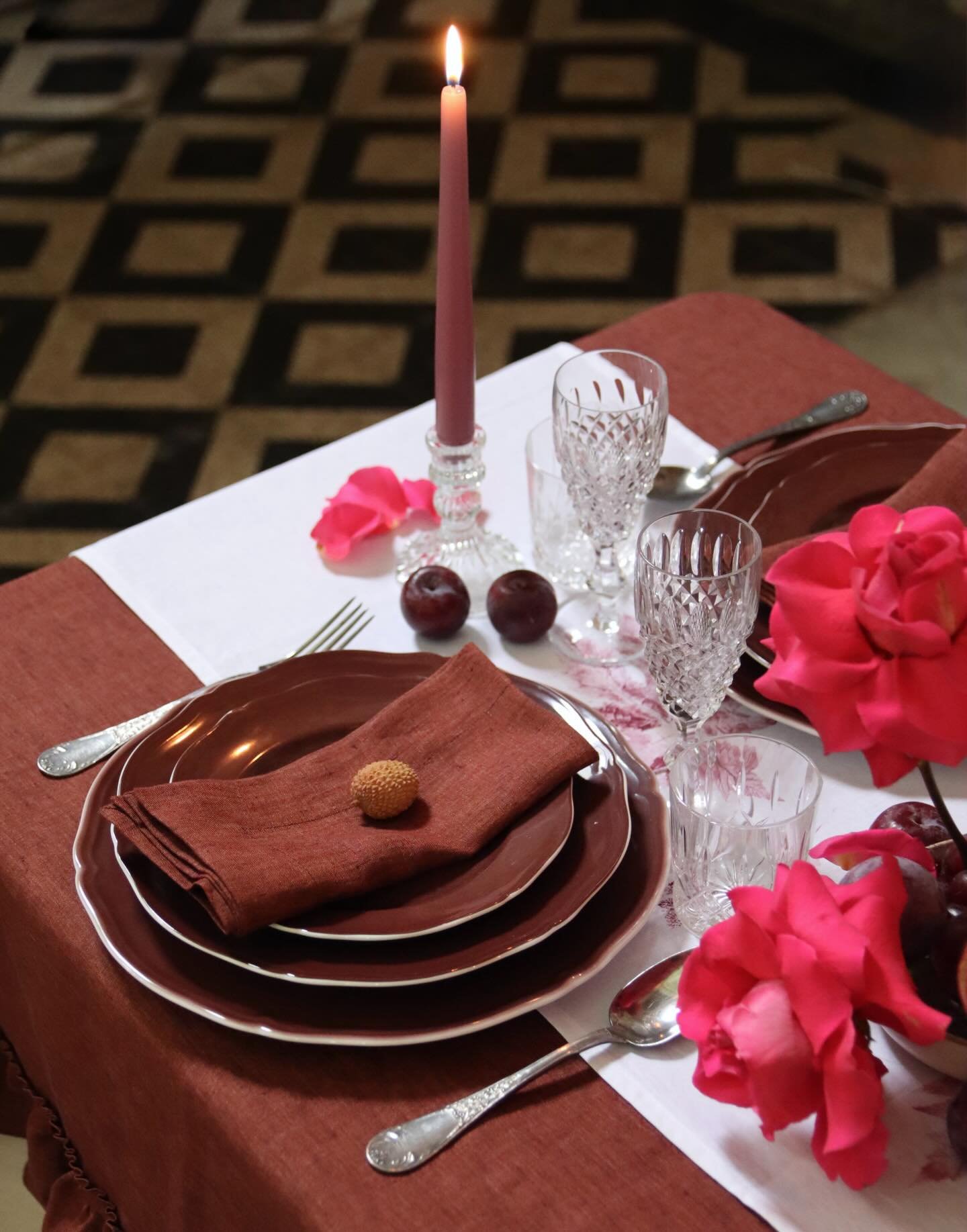 Tavolo apparecchiato con piatti, bicchieri di cristallo, posate, una candela rosa accesa, una fiore rosa, e due fichi su un tovagliolo di tessuto marrone e un centrino bianco, in un ambiente elegante.