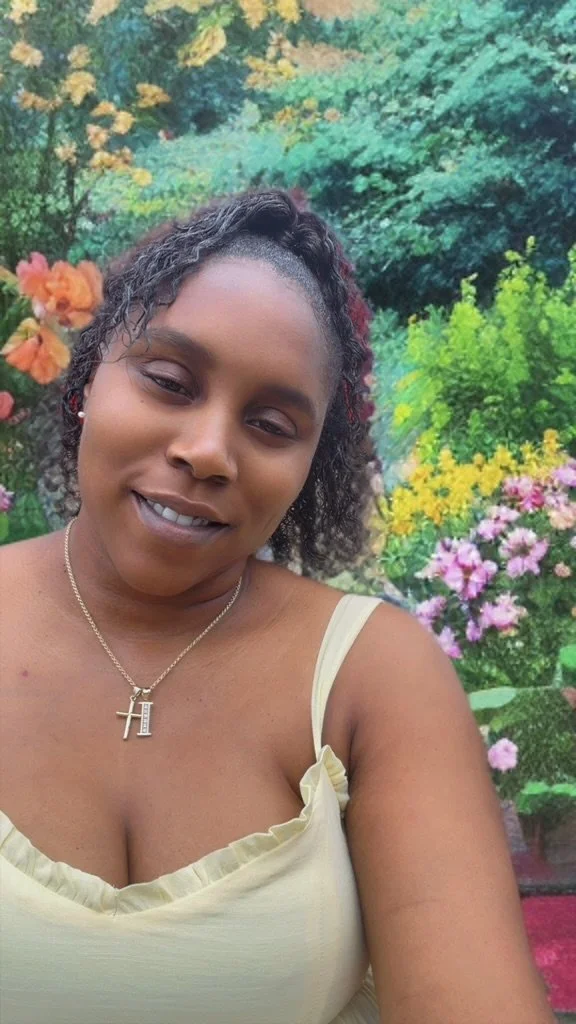 A woman with dark curly hair, wearing a yellow tank top and a gold necklace with an 'H' pendant, is smiling slightly while taking a selfie outdoors among colorful flowers and green foliage.
