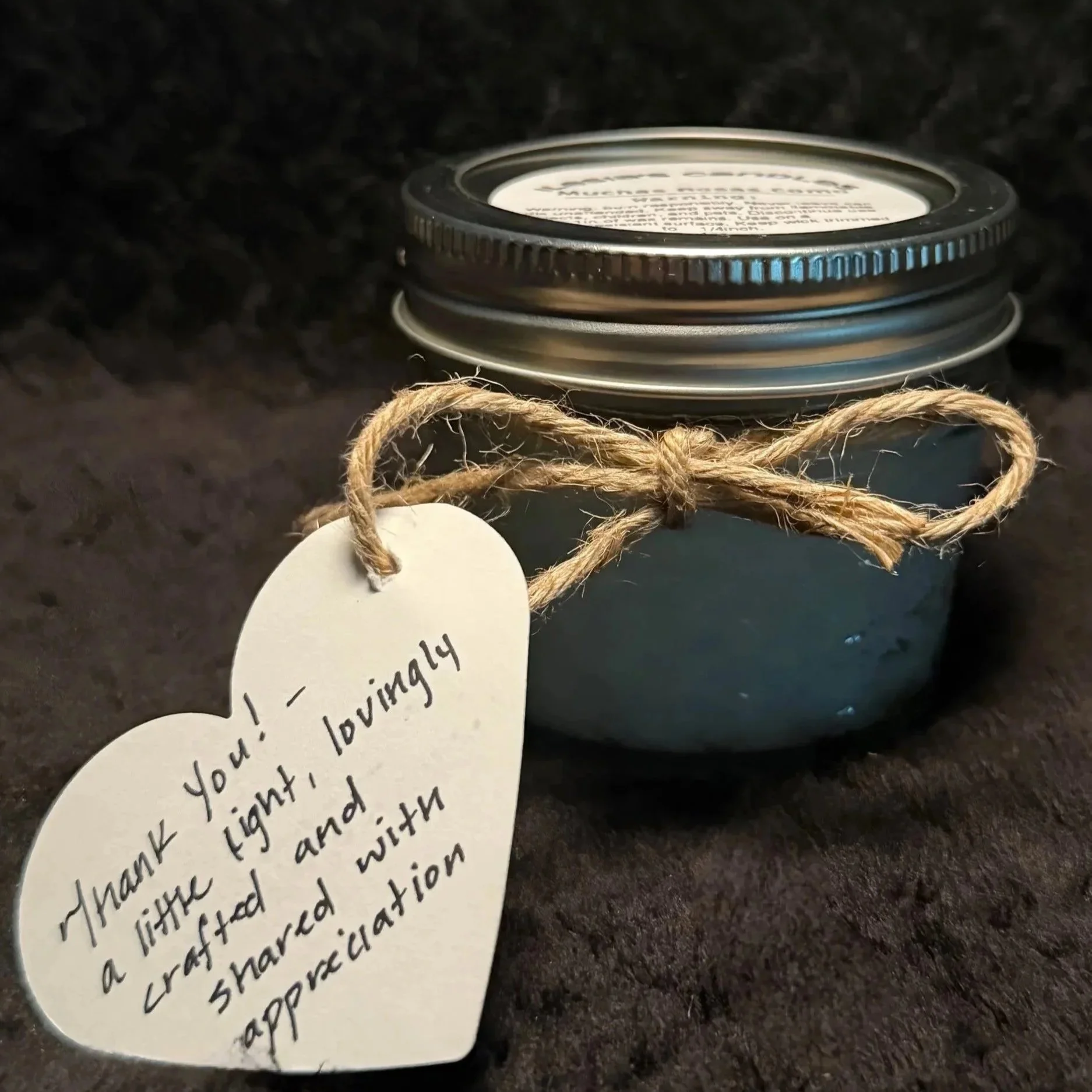 Close-up of a small jar with a metal lid wrapped with a jute twine bow, and a handwritten white paper gift tag attached to it. The jar is placed on a dark surface.