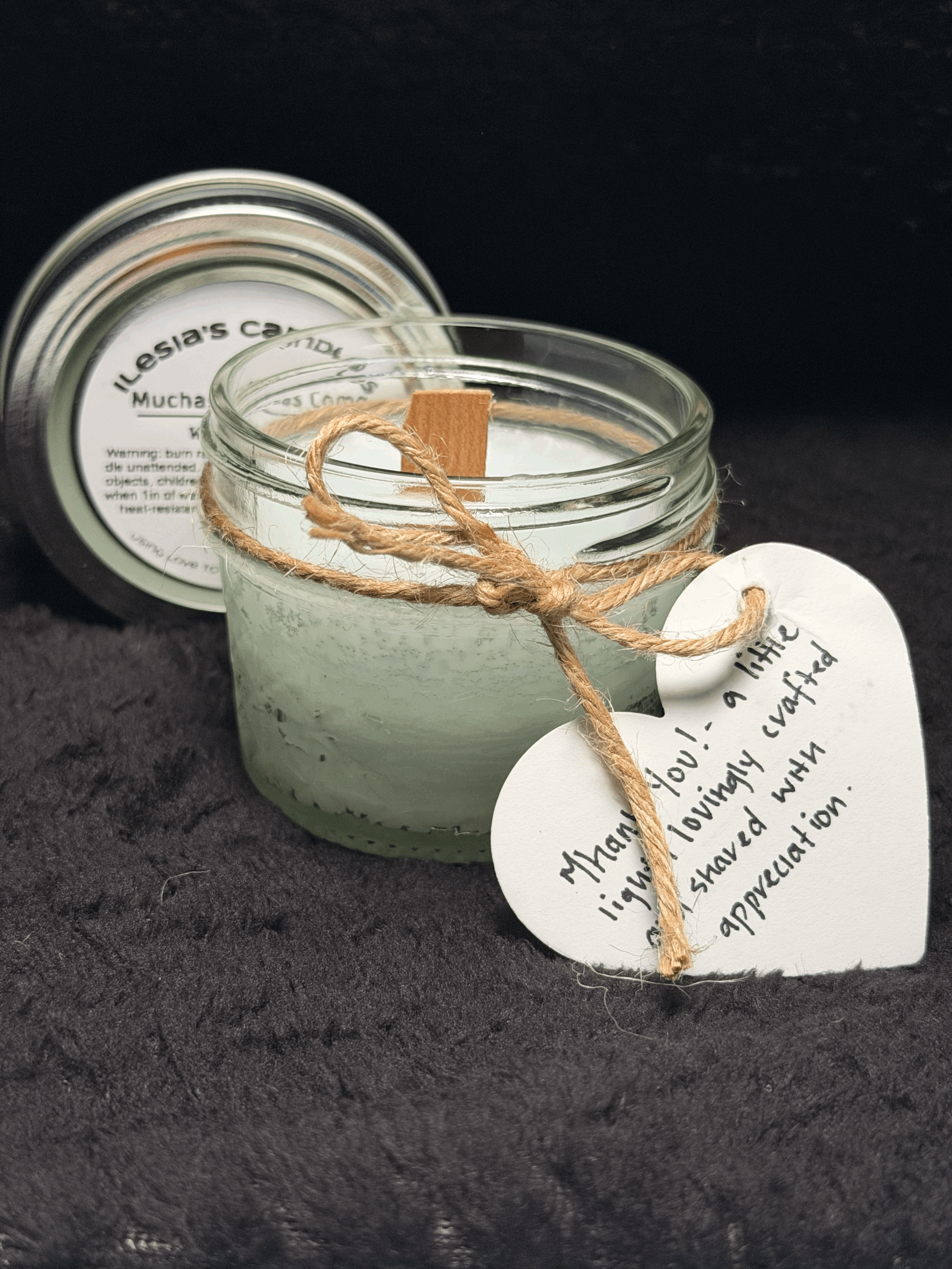 A white candle in a glass jar tied with a brown string and a small white tag with handwritten note, placed on a dark surface. The candle's lid is nearby, and the jar has a label with text partially visible.