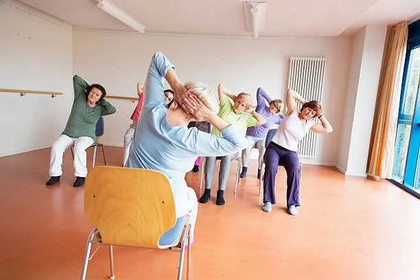 Group of seniors participating in a seated stretching class in a bright room with large windows and yellow curtains.