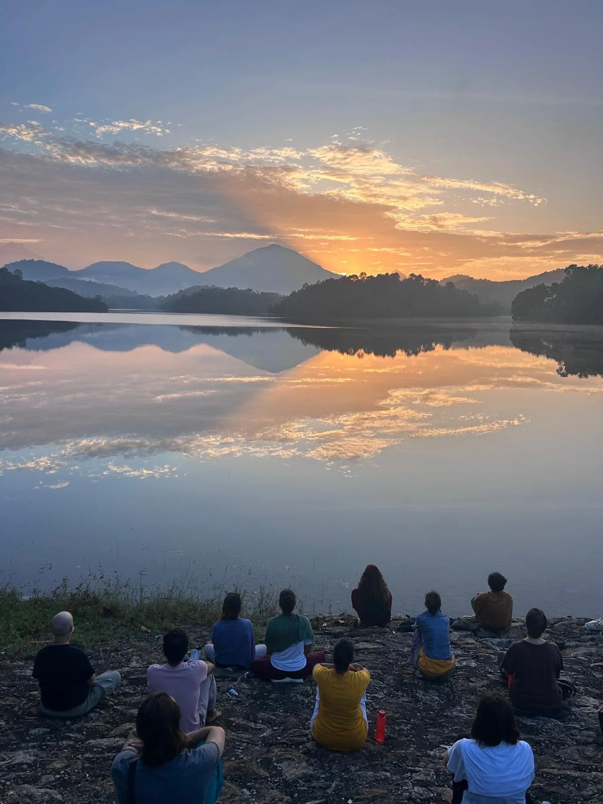 Group of people sitting on rocks by a lake watching a sunset with mountains in the background