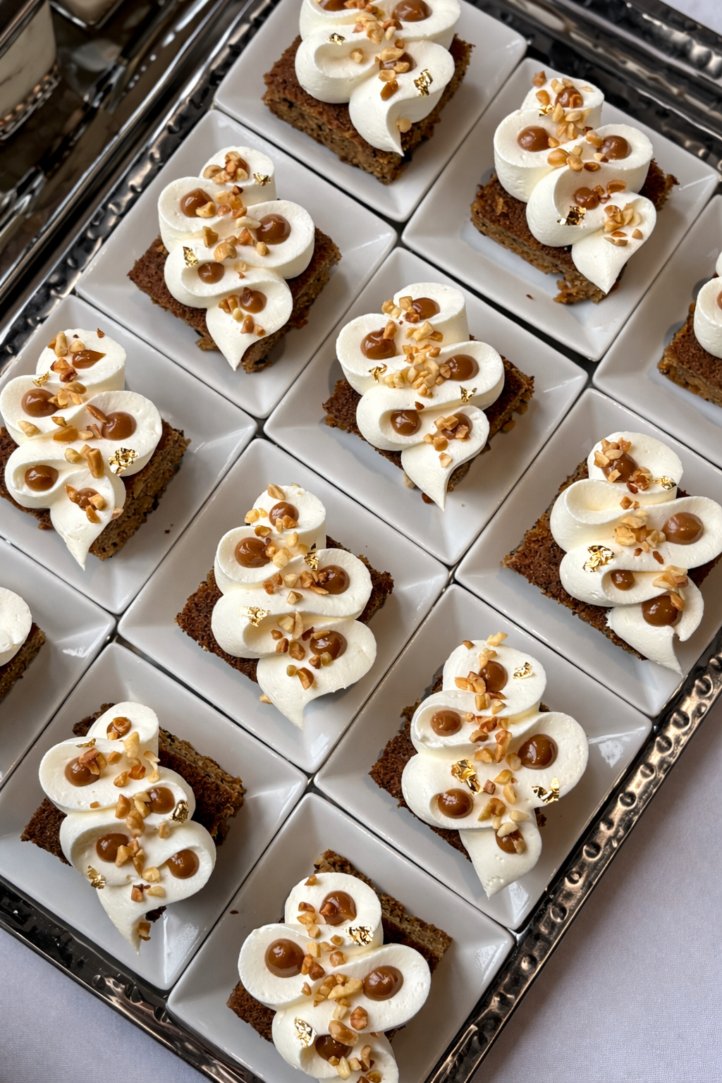 Set of small square chocolate cakes topped with whipped cream, caramel drizzle, chopped nuts, and gold leaf, arranged on a silver tray.