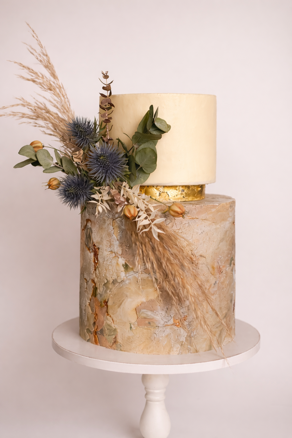 Two-tiered wedding cake with a textured marble pattern, decorated with dried flowers, leaves, and grasses, on a white cake stand.