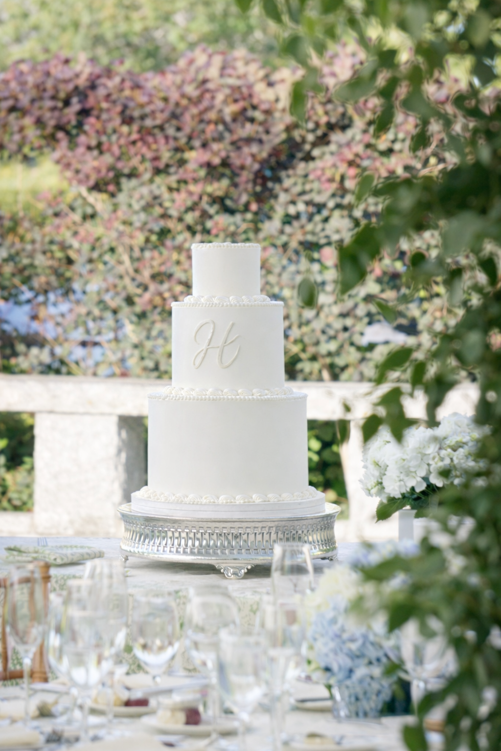 A white, three-tiered wedding cake with a letter 'H' on the middle tier, displayed on a silver cake stand outdoors with trees and flowers in the background.