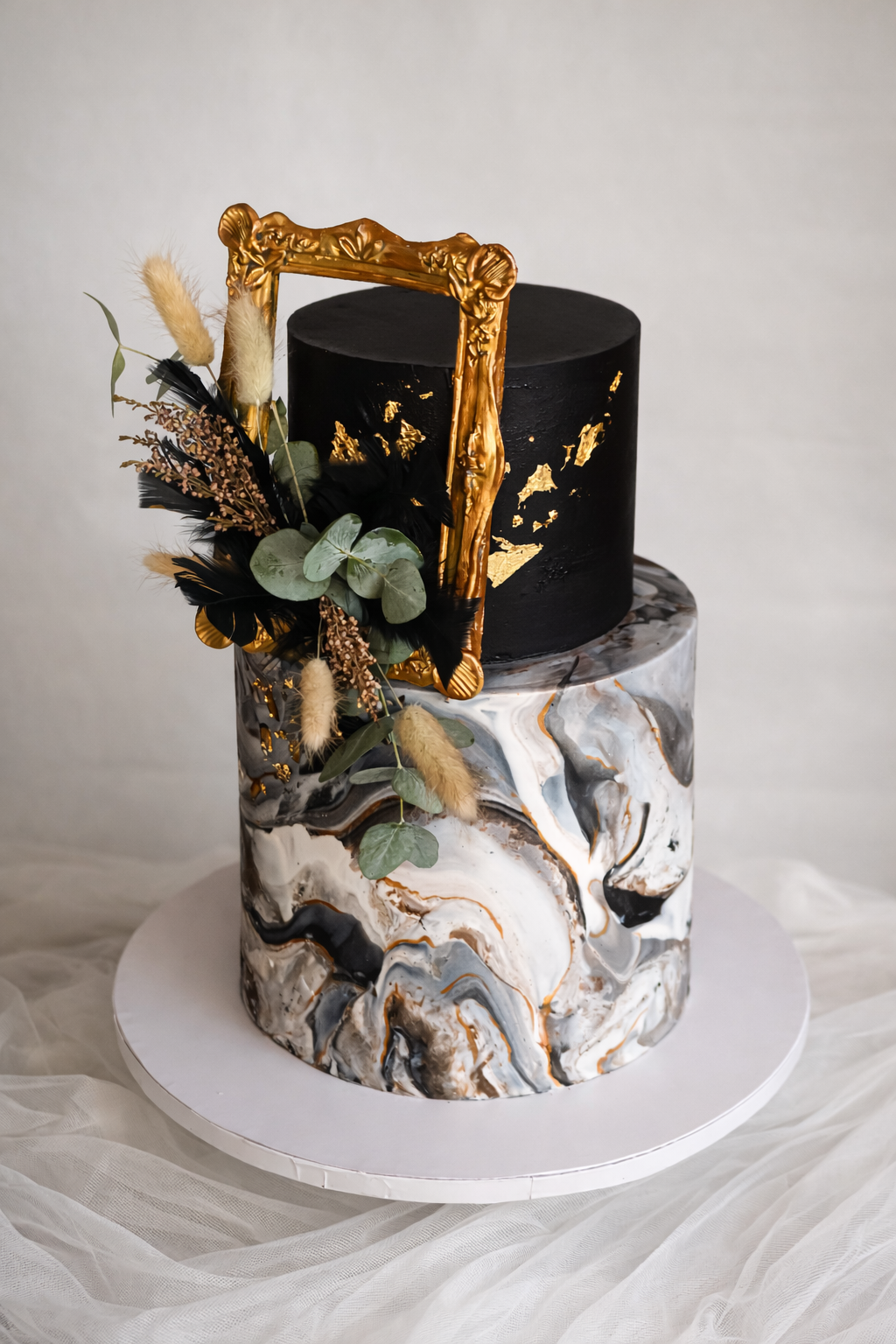 Two-tiered cake with marbled black, white, and gold pattern on the bottom layer and black with gold accents on the top layer. Decorated with a gold picture frame, dried flowers, eucalyptus leaves, and black feathers.