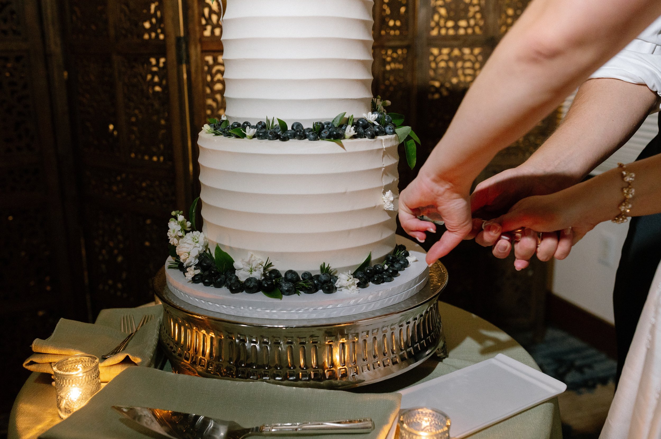 A bride and groom cutting a wedding cake decorated with blueberries, white flowers, and green leaves, on a gold cake stand, at a wedding reception.
