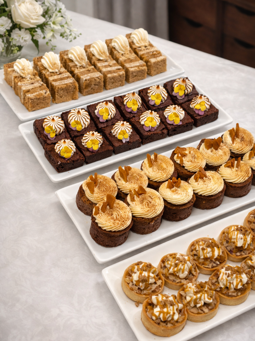 Assorted mini desserts on white rectangular plates, including coffee-flavored squares with whipped topping, chocolate brownies with floral decorations, caramel-topped mini cheesecakes, and caramel-topped tarts filled with nut and cream ingredients, on a marble table with a floral centerpiece in the background.