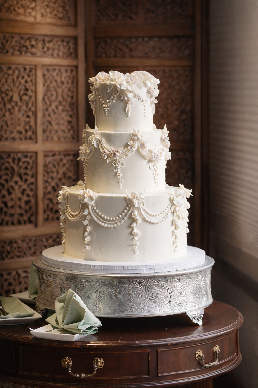 A tall three-tier wedding cake with white icing and decorative sugar piping, flowers, and pearl details, placed on an ornate silver cake stand on a wooden table.