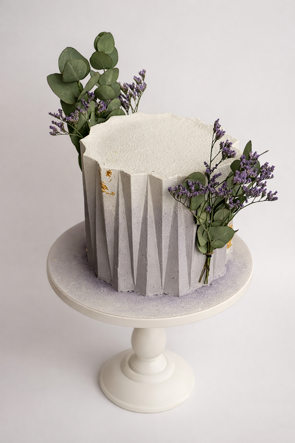 A white textured cake with a geometric design around the sides, decorated with purple flowers and green leaves, on a white cake stand against a plain white background.
