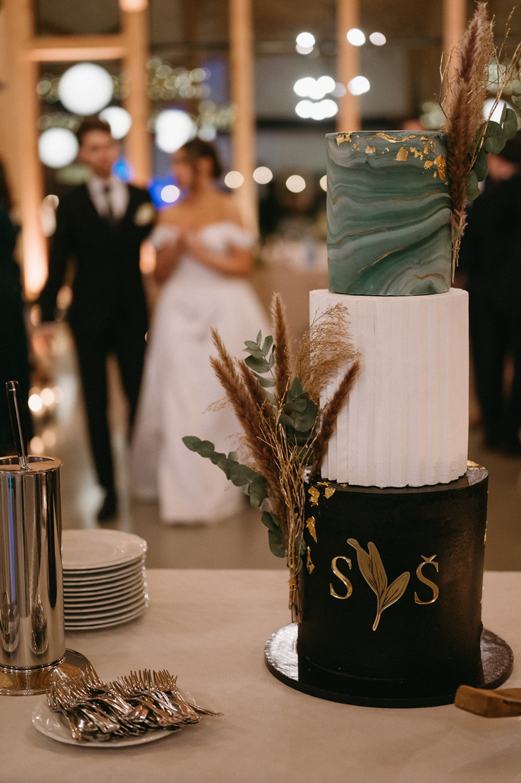 Wedding cake with three tiers, decorated with greenery and pampas grass, featuring a black bottom tier with gold letters and a green marbled top tier, on a table with plates and silverware. Blurred couple in wedding attire in the background.