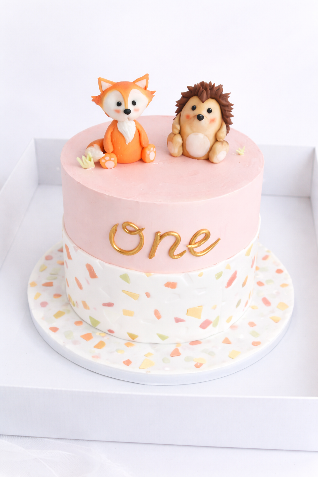 A two-tiered birthday cake with pink and white icing decorated with sprinkles, topped with cute animal figures of a fox and a hedgehog, and the word 'One' written in gold.