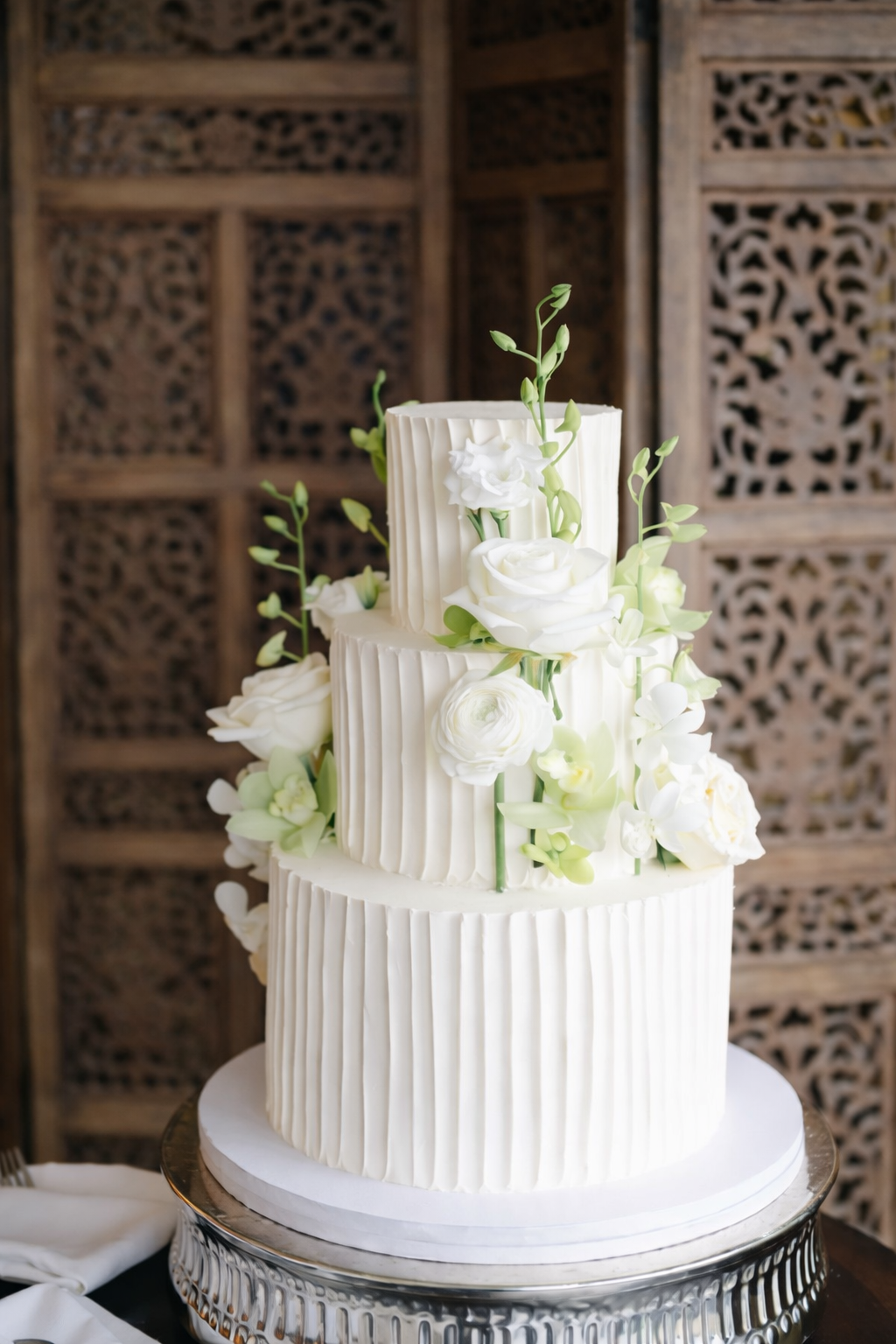 A three-tiered white wedding cake decorated with white flowers and green foliage, featuring vertical ridges on the sides, on a silver cake stand.