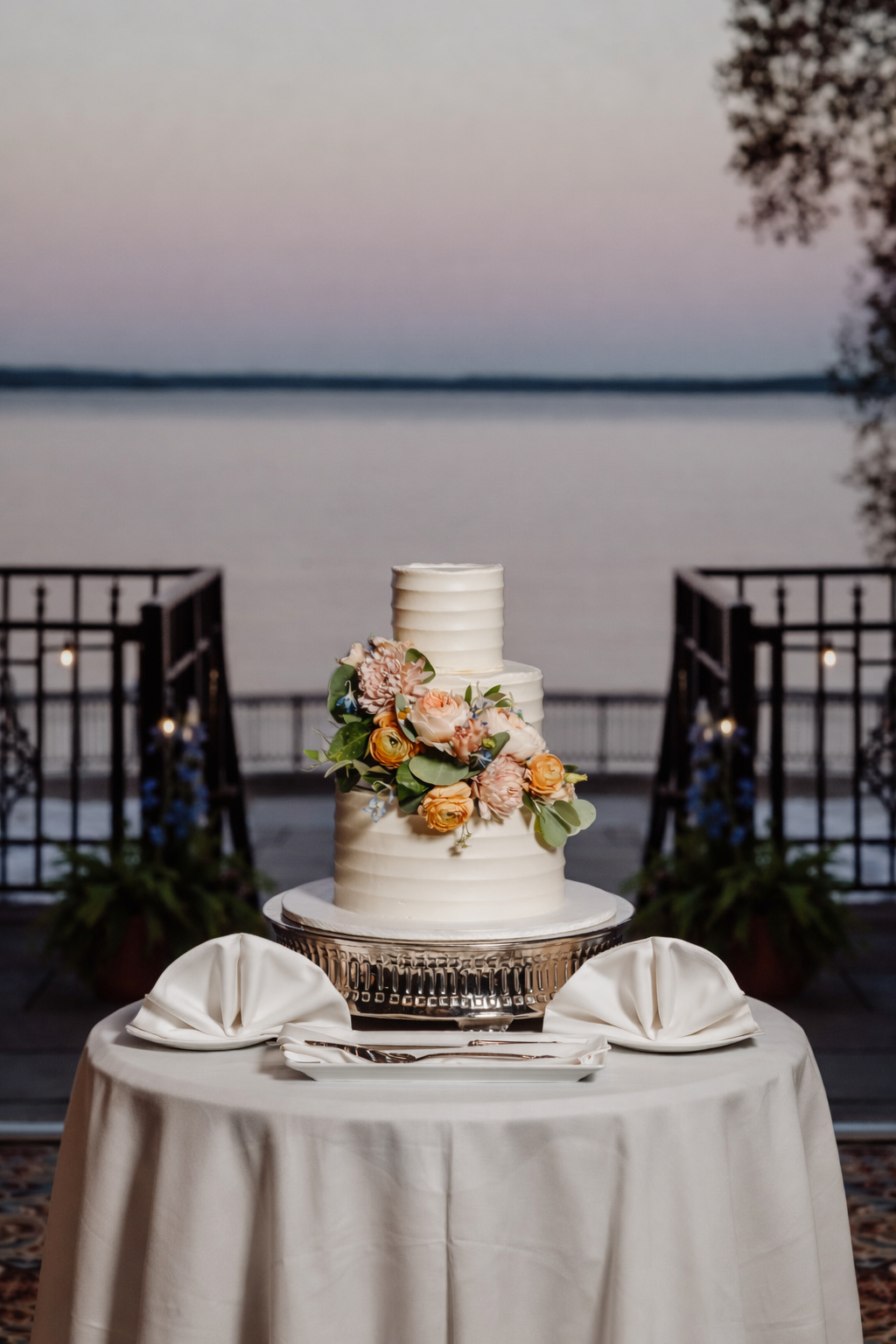 Elegant two-tier white wedding cake decorated with a pink and orange floral arrangement, placed on a table with white cloth and silver cake stand overlooking a lake at dusk.
