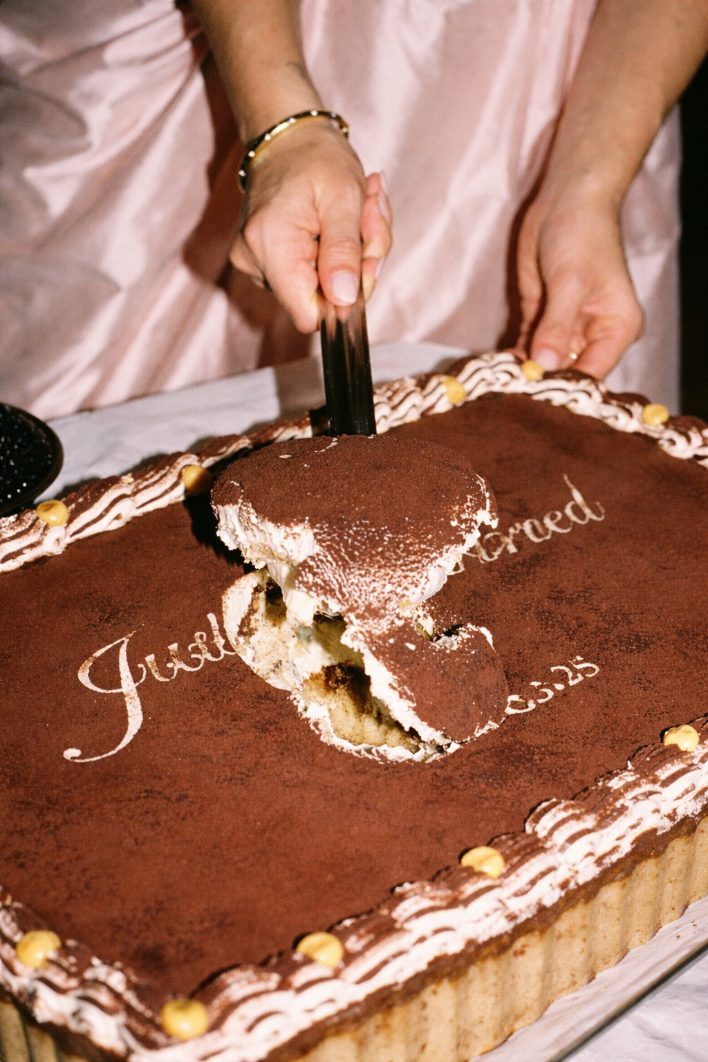 Person in pink dress slicing a large rectangular chocolate cake with a cake server.