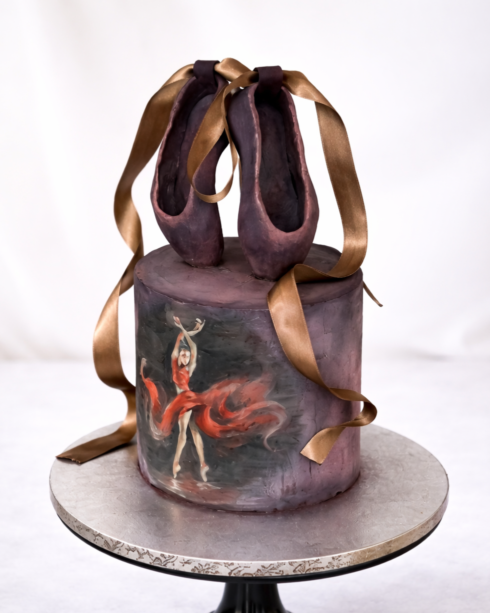 A decorative cake with ballet-themed artwork, featuring ballet slippers on top tied with satin ribbons, and a painted ballerina with a red flowing dress on the side of the cake.