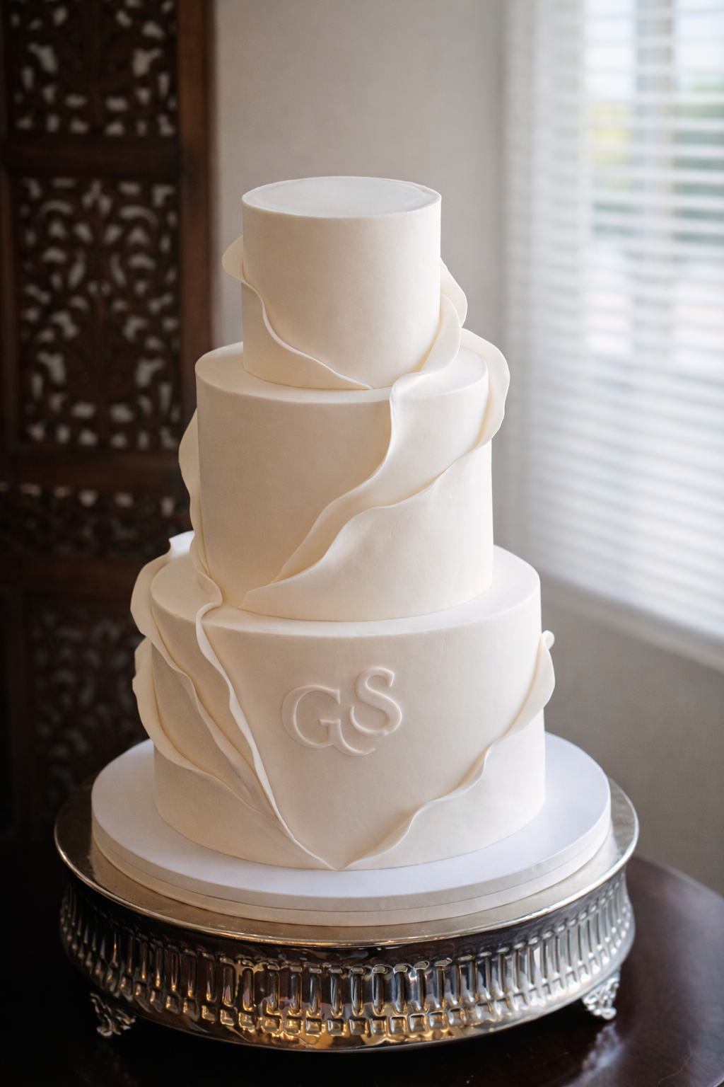 A four-tier white wedding cake with decorative white fondant ribbons, featuring the initials 'GS' on the bottom tier, placed on a silver cake stand near a window with white blinds.