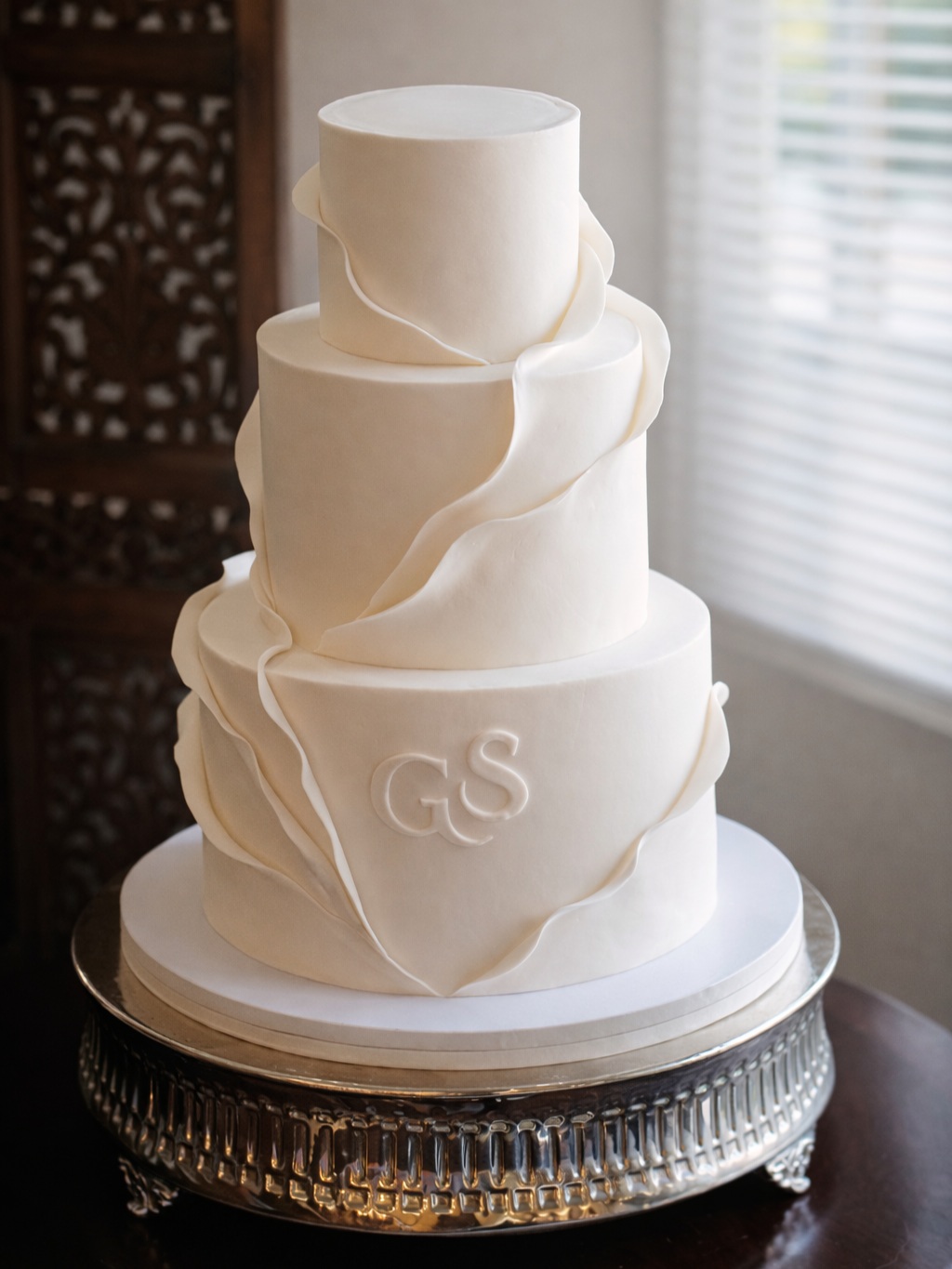 A four-tier white wedding cake with elegant wavy decorations and the initials "GS" on the lower tier, placed on a silver cake stand.
