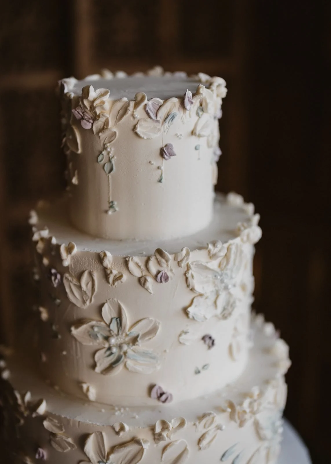 Three-tiered white wedding cake decorated with piped buttercream flowers and petals, set against a dark background.