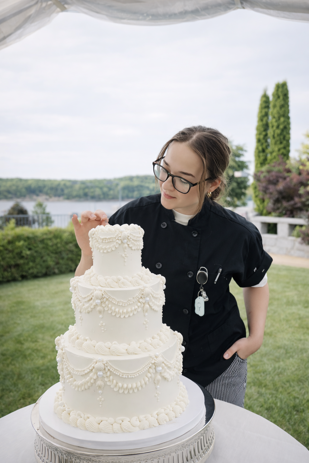 A woman in a black chef's uniform and glasses stands outdoors, looking at an elaborately decorated, tiered white cake on a table. The cake features intricate piping and decorative elements, with a river and trees visible in the background under a par