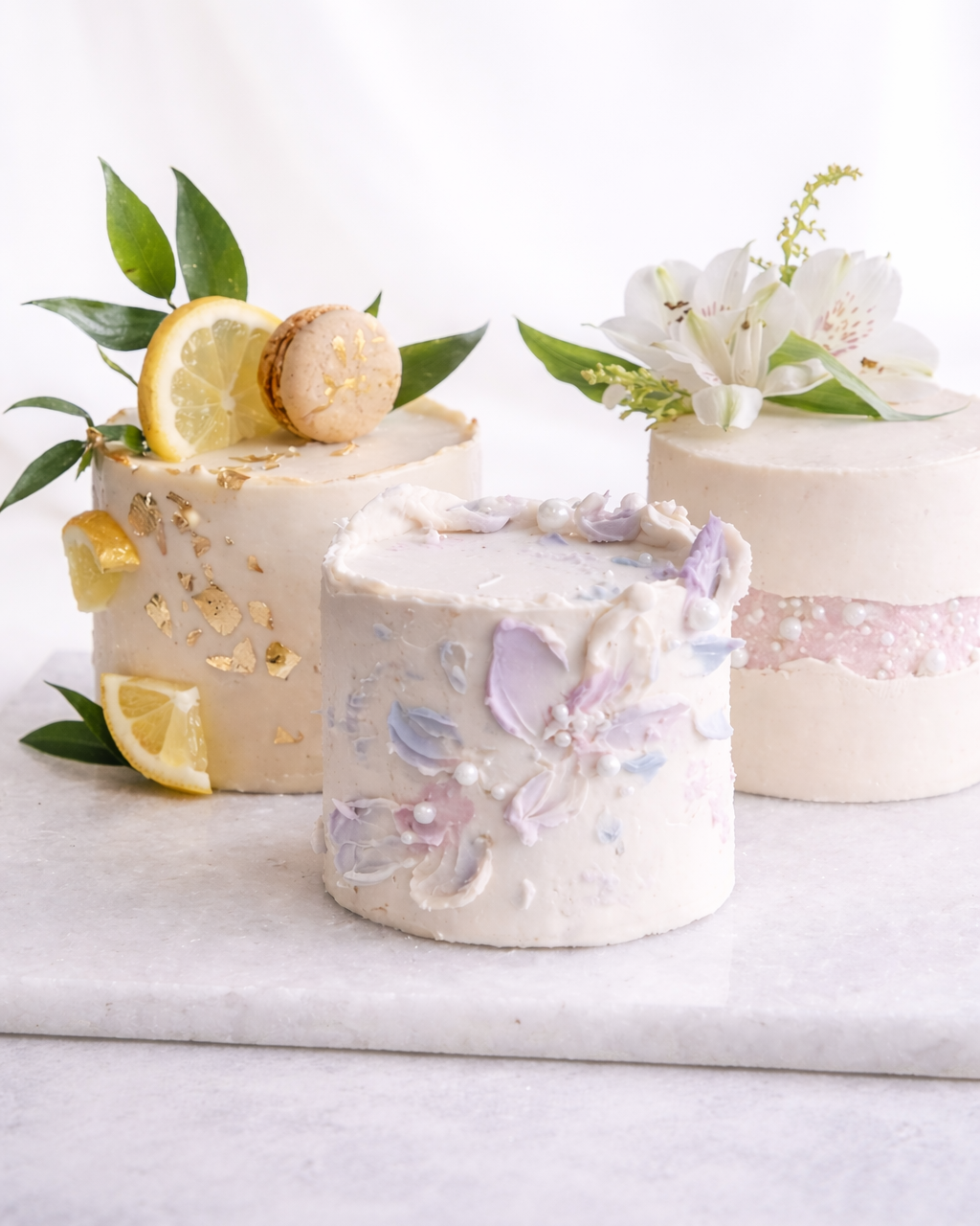 Three elegant wedding cakes decorated with flowers, lemon slices, and macarons, against a white background.