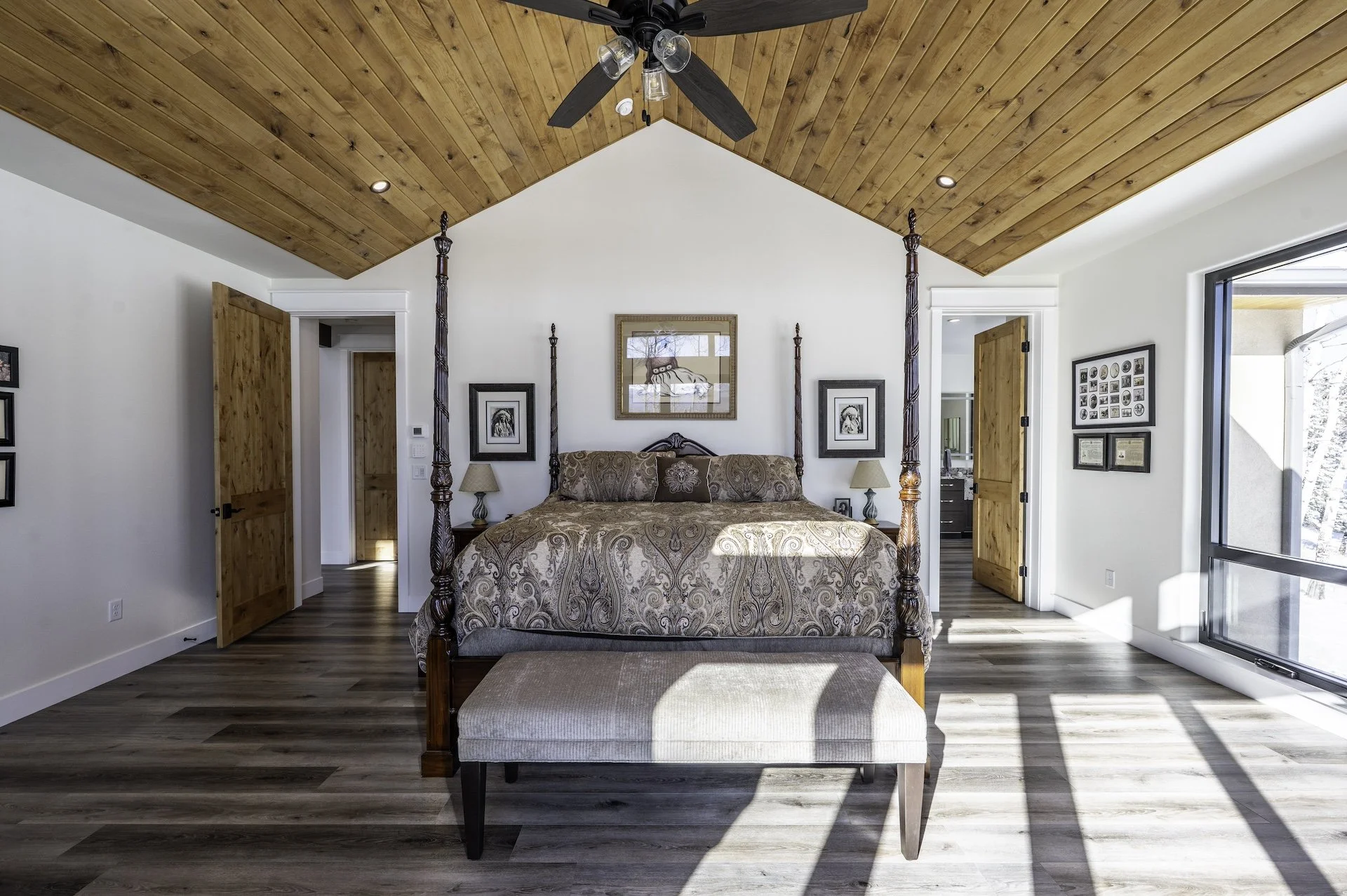Bright bedroom with a four-poster bed, patterned bedding, and a wooden vaulted ceiling. Sunlight streams through a large window.