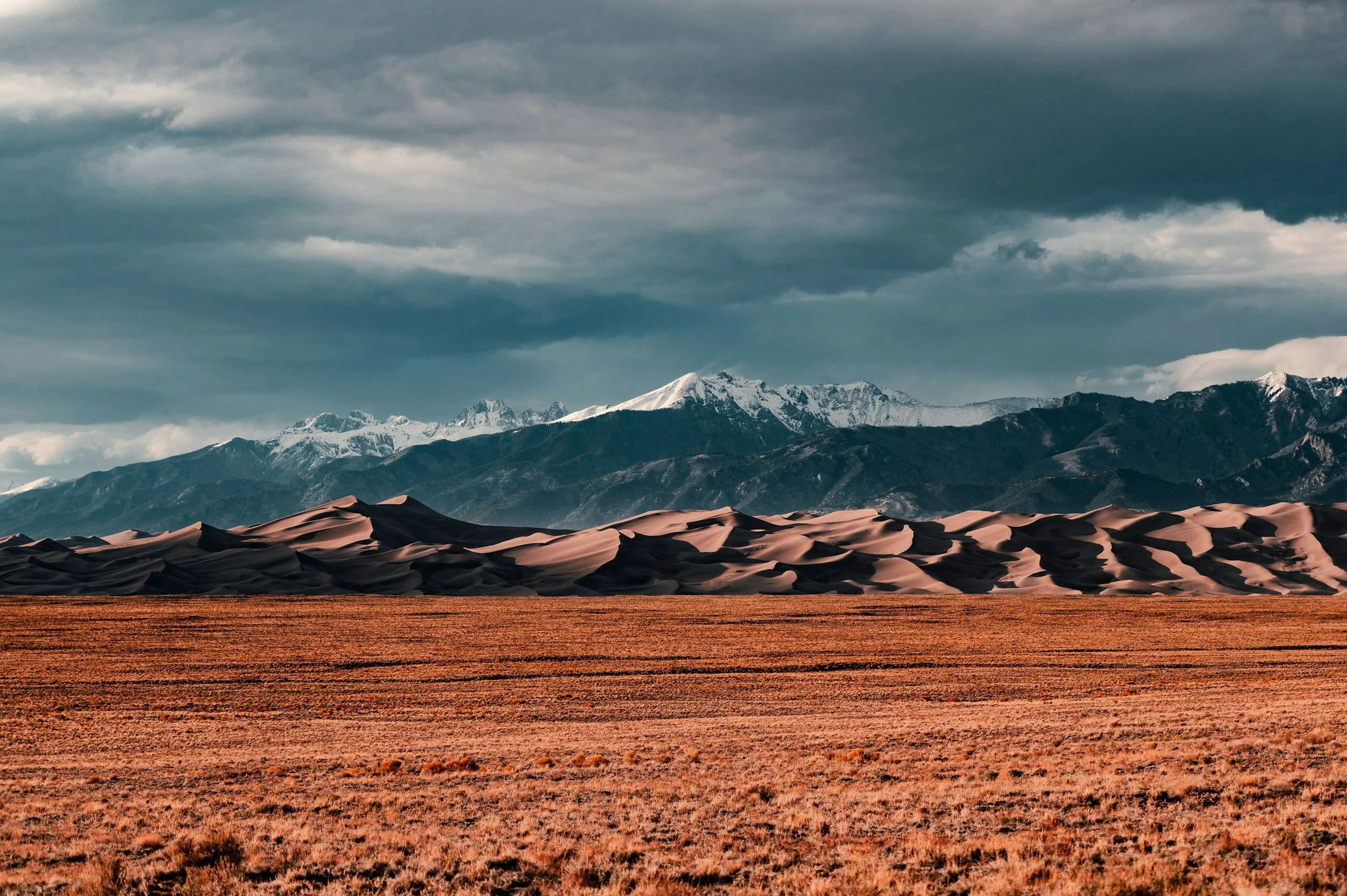 A desert landscape with sand dunes in the foreground, mountain range with snow-capped peaks in the background, and a cloudy sky overhead.