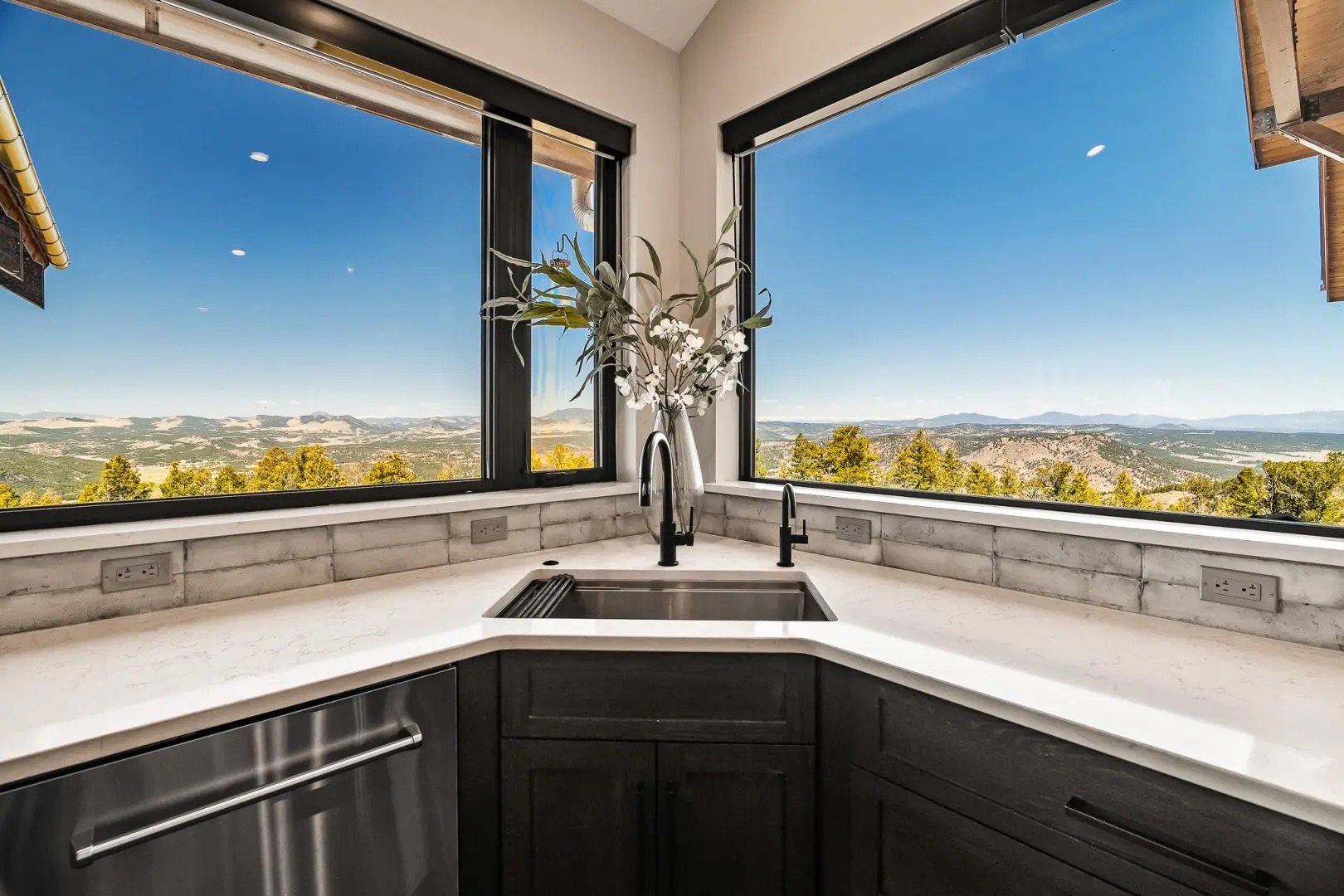 Kitchen with a large window view of mountains and trees, a white marble countertop, a stainless steel sink, black faucet, and a vase with flowers on the window sill.