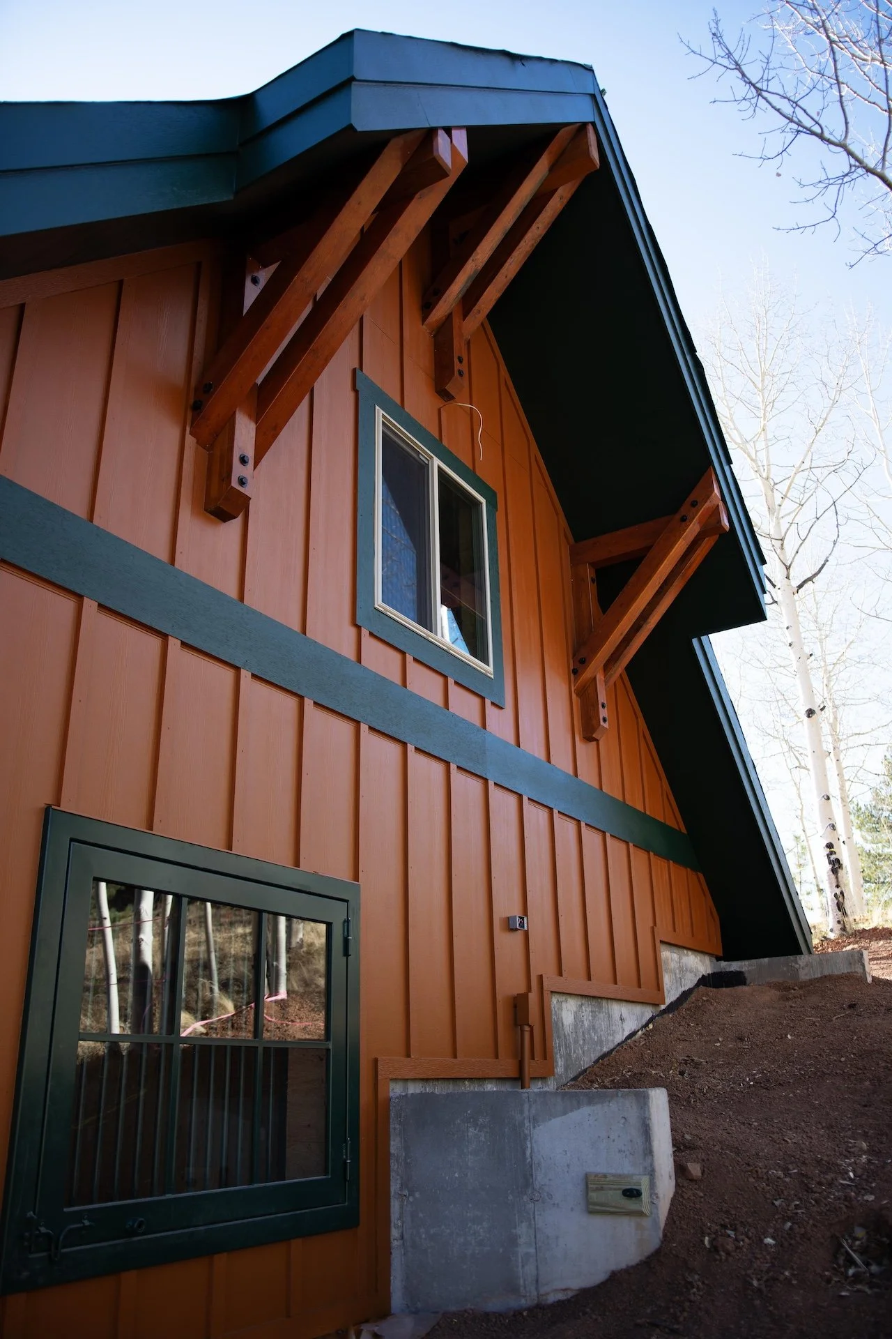 Close-up of a wooden house exterior showing a window, green trim, and metal supports for the roof overhang, with a sloped dirt ground and bare trees in the background.