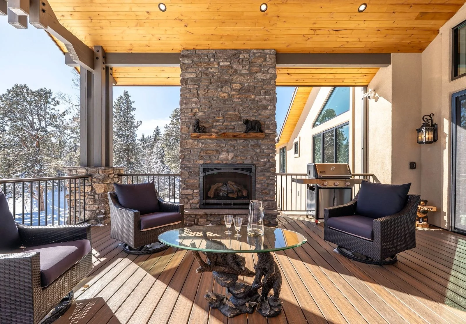 A cozy outdoor patio with wooden flooring, a stone fireplace, and seating area including wicker chairs with purple cushions. There is a glass-top table with a decorative wooden base, a grill, and snow-covered trees visible in the background through the open railing. The ceiling is wood-paneled with recessed lighting, and a lantern-style light fixture is mounted on the wall.