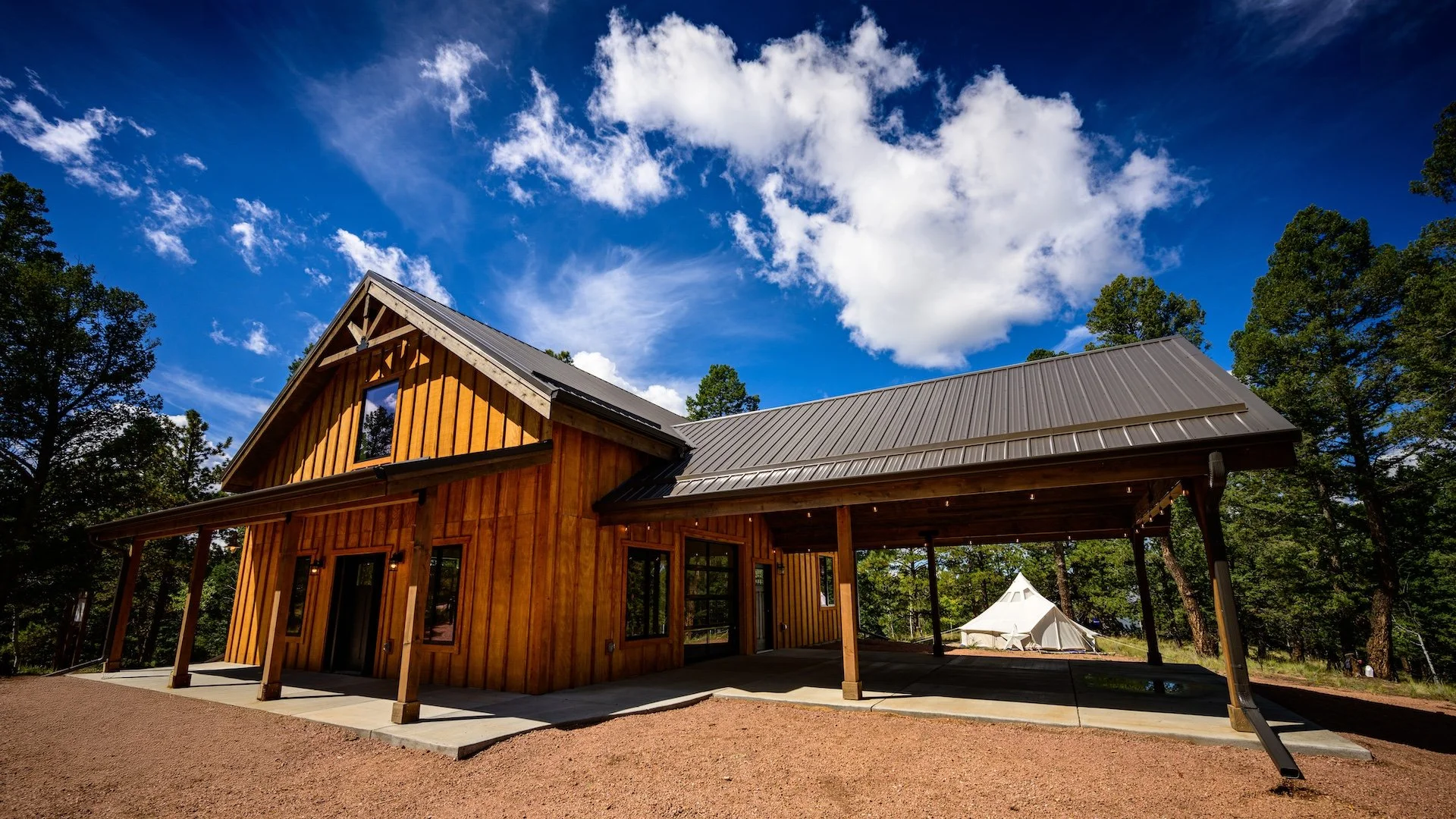 A two-story wooden house with a metal roof, surrounded by trees and under a blue sky with clouds, with a porch and a covered carport.