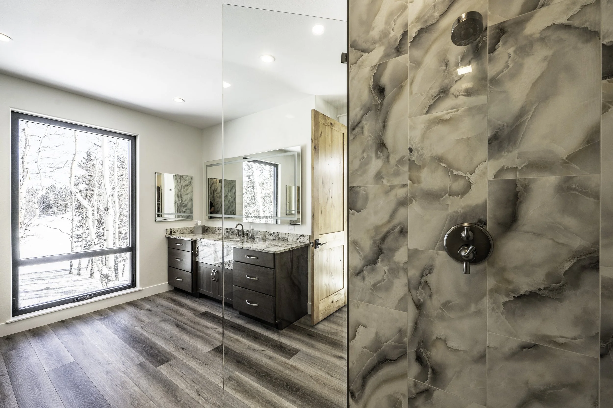 A modern bathroom with a large window showing snow-covered trees outside, featuring a dual-sink vanity with granite countertops, dark wood cabinets, a large mirror, and a marble-tiled shower area with a rain showerhead.