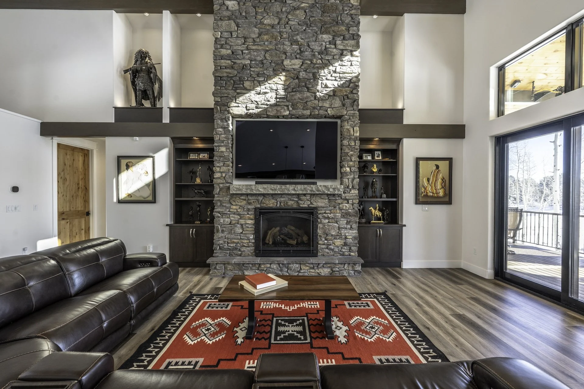 Living room with a stone fireplace, a wall-mounted TV, bookshelves with figurines, a leather sofa, a wooden coffee table with books, a red and black patterned rug, and sliding glass doors to an outdoor balcony.
