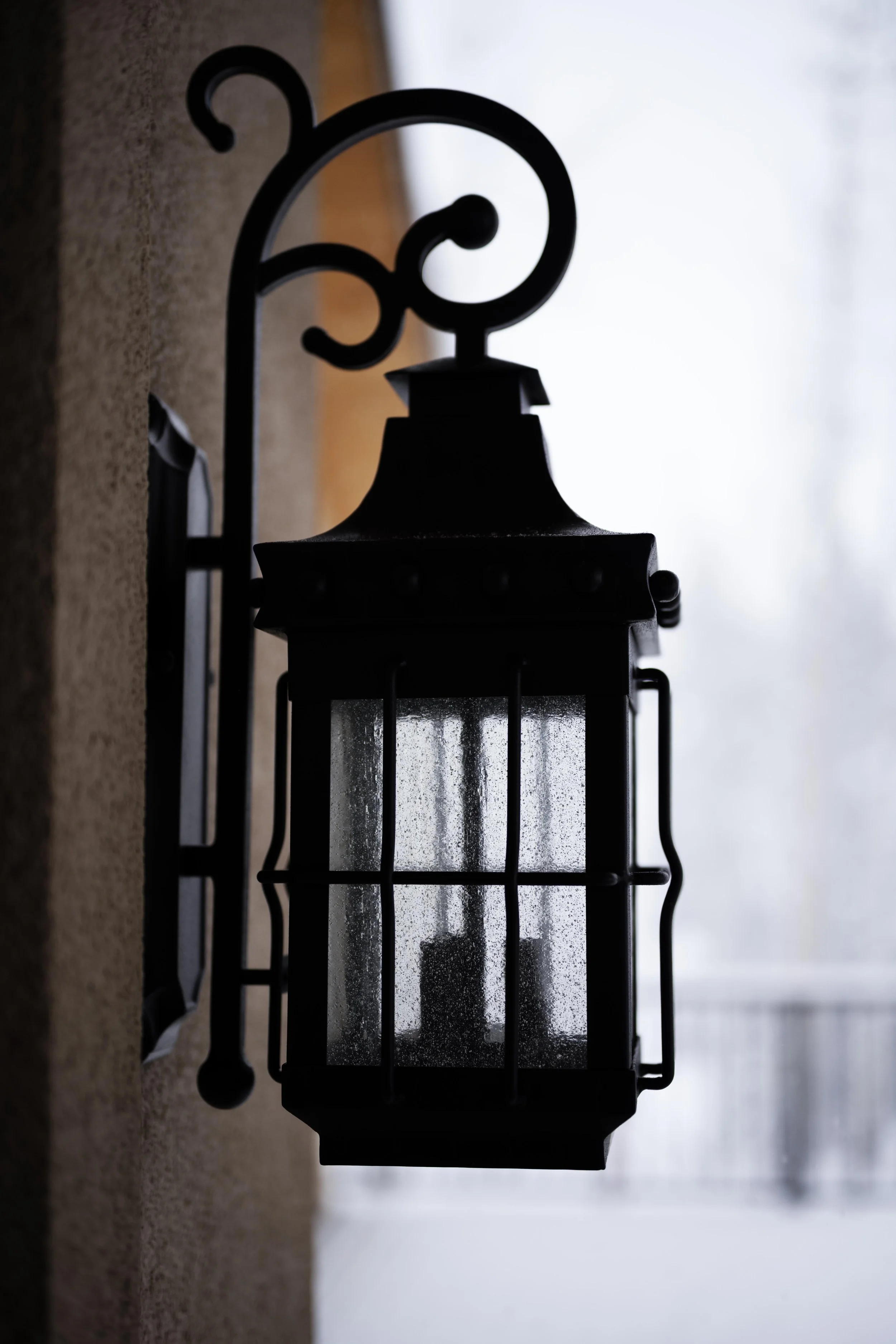 Black outdoor wall lantern with rain droplets on glass, mounted on a beige wall, with a blurred background of a snowy or foggy outdoor scene.
