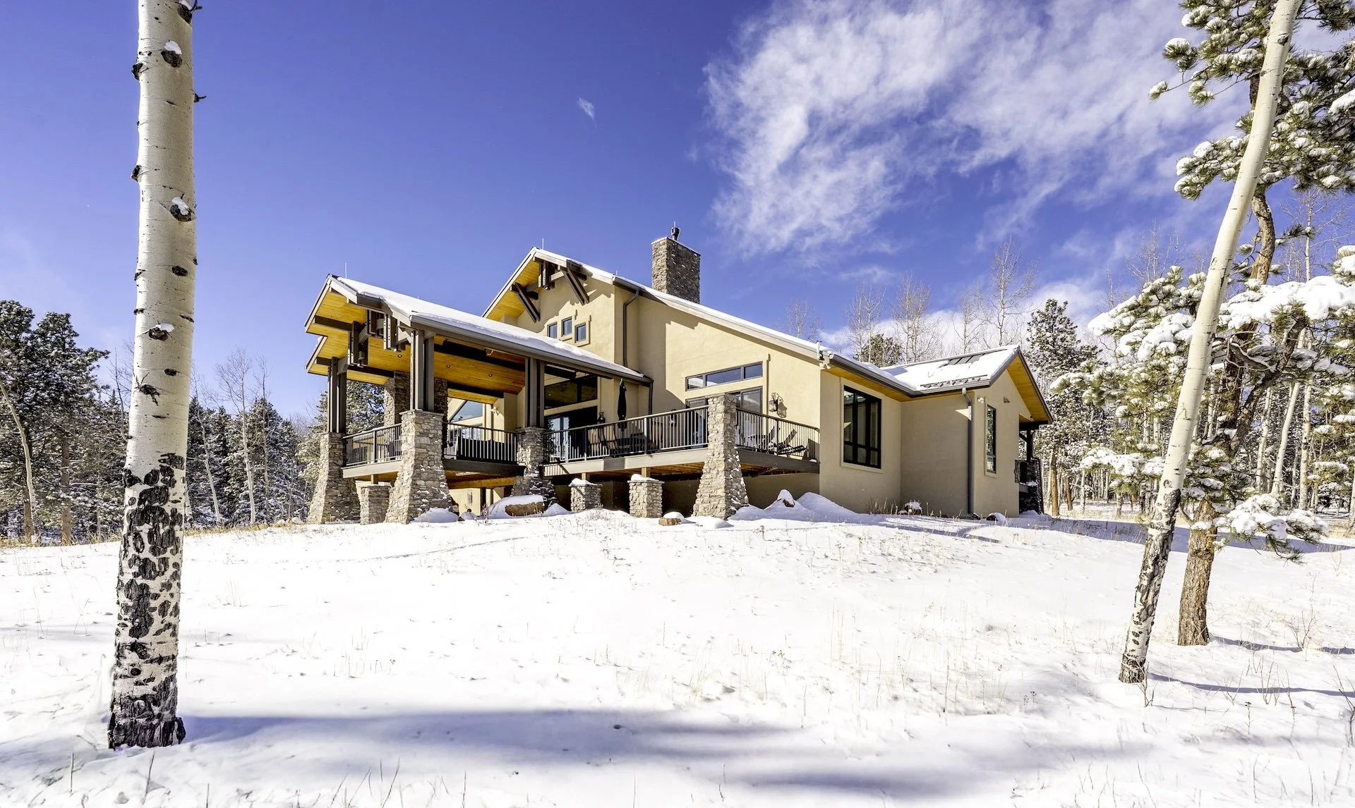 A modern house with a beige exterior, stone columns, and large windows in a snowy landscape with trees and a clear blue sky.
