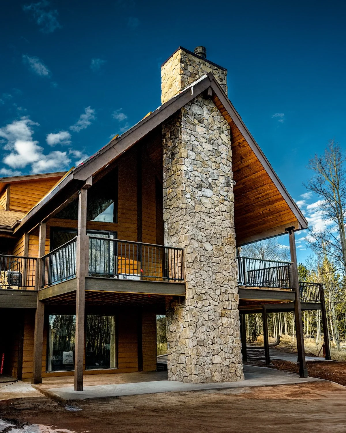 A modern two-story house with a large stone chimney, wooden siding, and an outdoor balcony, set against a blue sky.