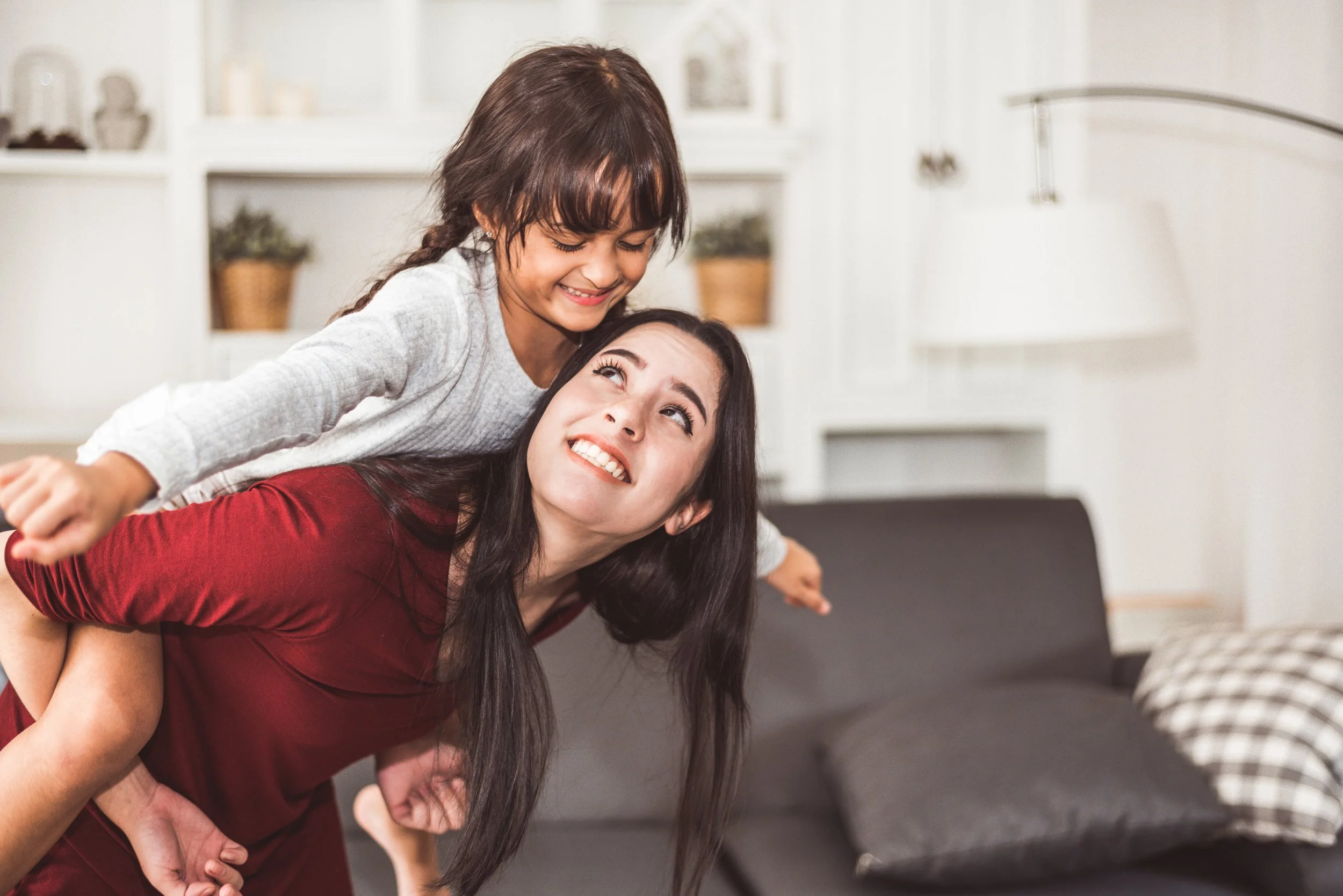 A young girl and a woman, likely mother and daughter, smiling and playing together in a living room, with the girl on the woman's back.