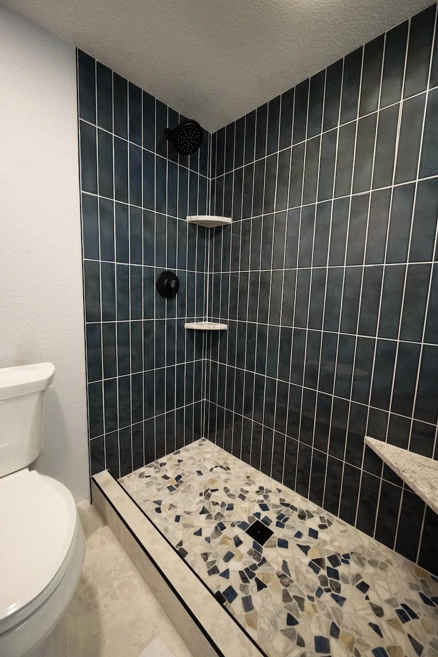 A modern walk-in shower with black tiled walls, a dark showerhead, black faucet, white and black mosaic tiles on the floor, and built-in marble shelves. A portion of a toilet is visible on the left.