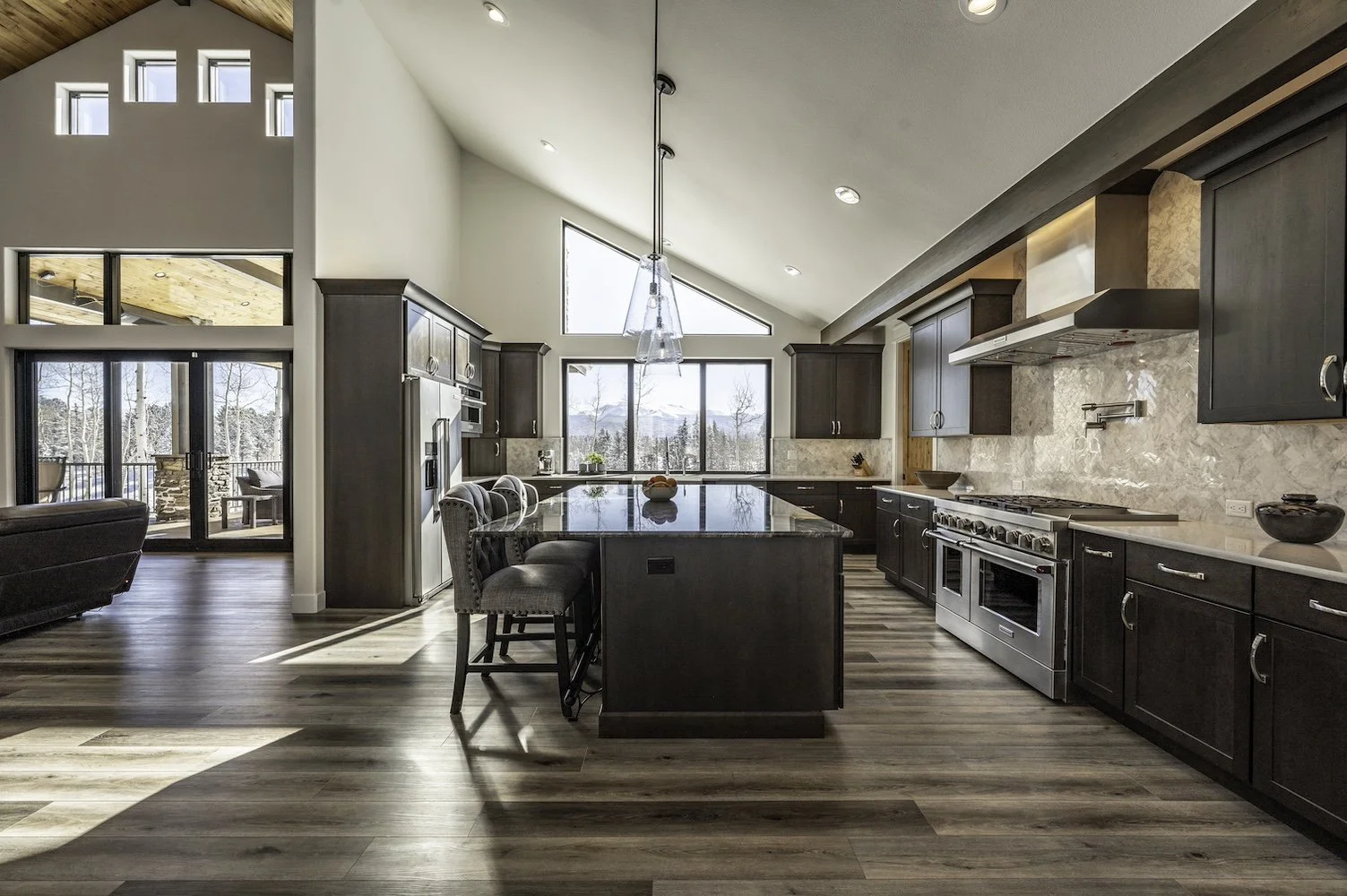 Modern kitchen with dark cabinets, a large island with seating, and view of mountains through large windows.