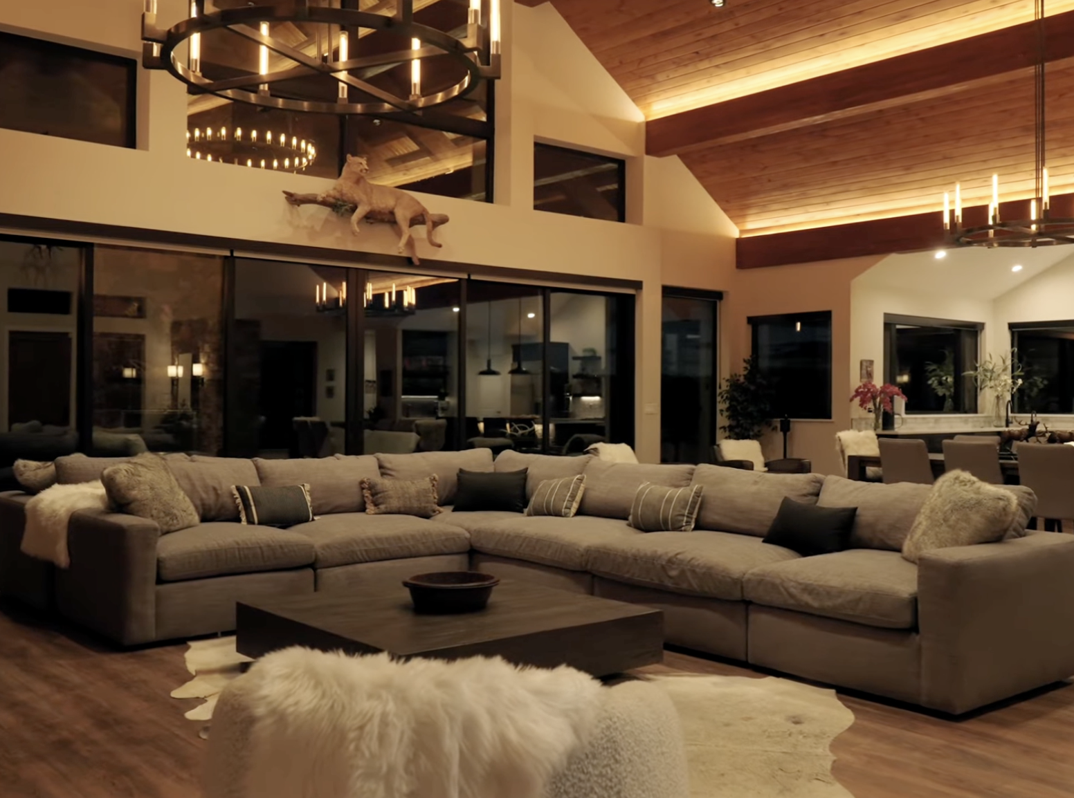 A spacious living room with a large L-shaped beige sectional sofa, multiple throw pillows, and a dark wooden coffee table. There's a white sheepskin rug in front of the sofa. The room features high ceilings with wooden paneling, large windows, and mo