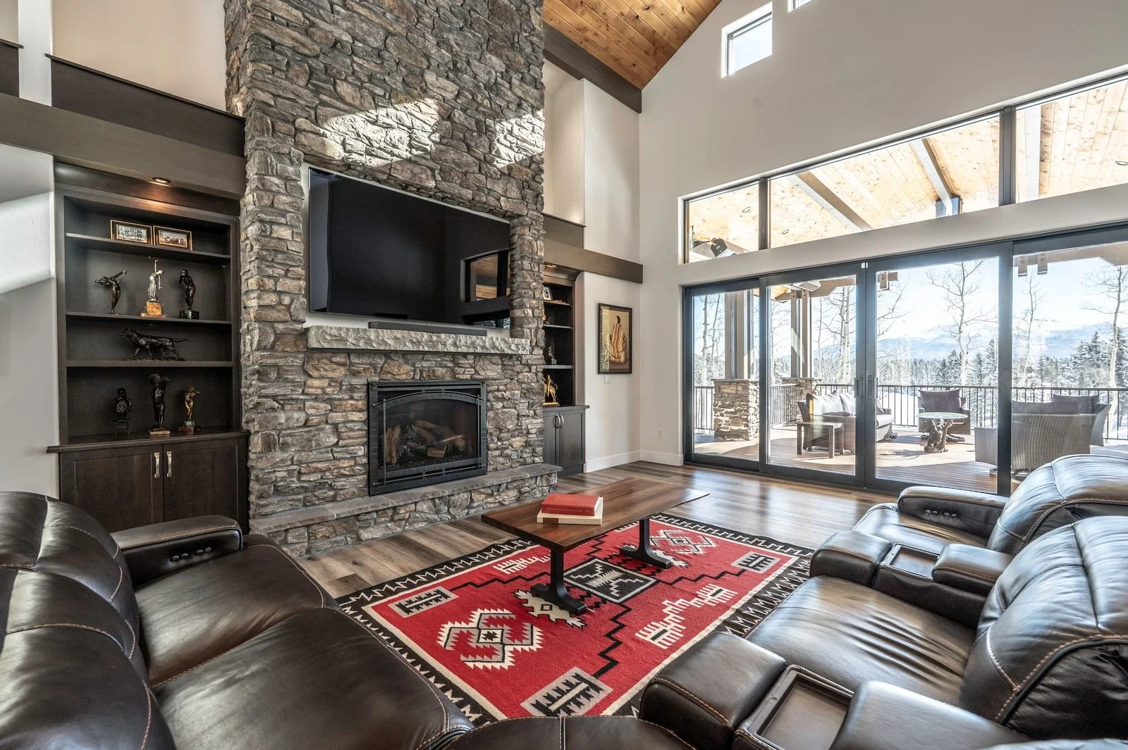 Living room with a stone fireplace, leather couches, a wooden coffee table with a book, a colorful patterned rug, and large glass sliding doors leading to a deck with outdoor furniture and a snowy landscape.