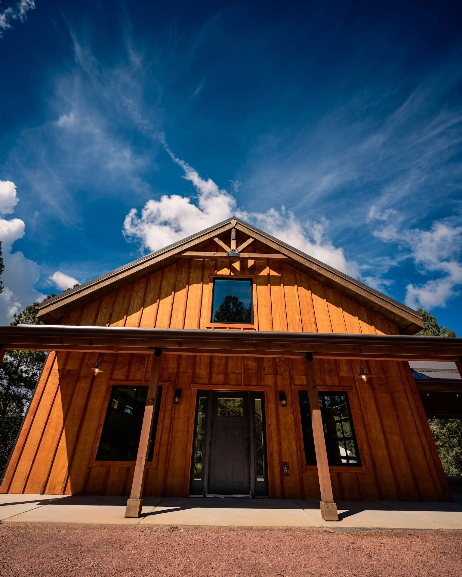 Front view of a modern wooden house with a gable roof, large windows, and a small porch under a bright blue sky with white clouds.
