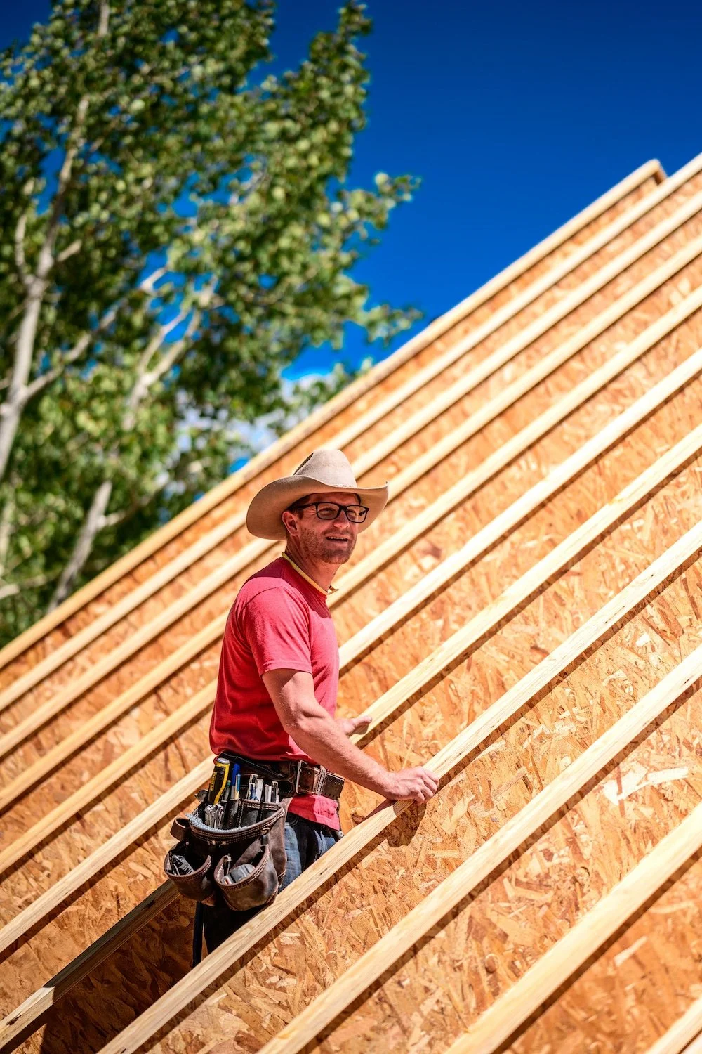 Colorado Custom Built owner Shobie Spear, wearing a cowboy hat, glasses, and a red t-shirt, standing on a construction site, holding onto wooden framing for a building with a clear blue sky and a tree in the background.