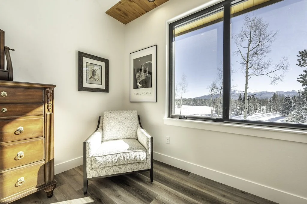 A cozy corner inside a room featuring a wooden dresser, a white armchair, and framed artwork on the wall. A large window reveals a snowy landscape with trees and distant mountains.