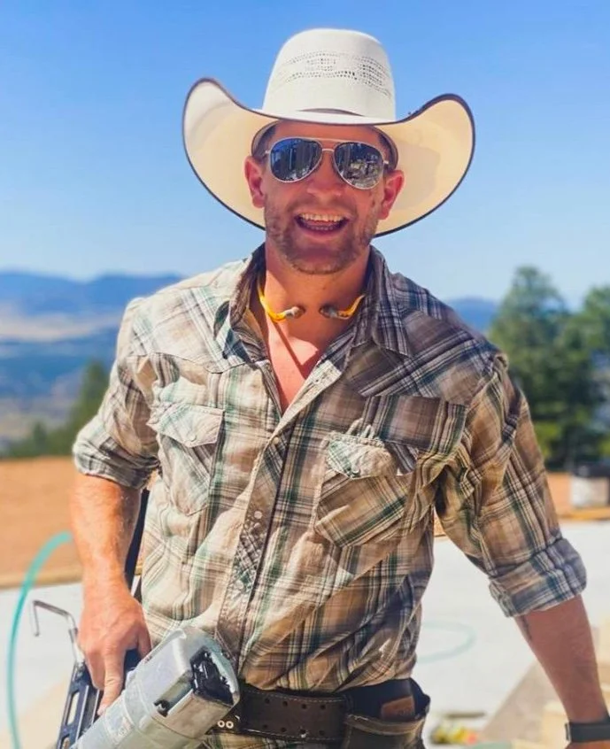 Shobie Spear, wearing a wide-brimmed cowboy hat, sunglasses, and a plaid shirt, smiles outdoors with mountains in the background. He is wearing yellow ear protection around his neck and holding a power drill.